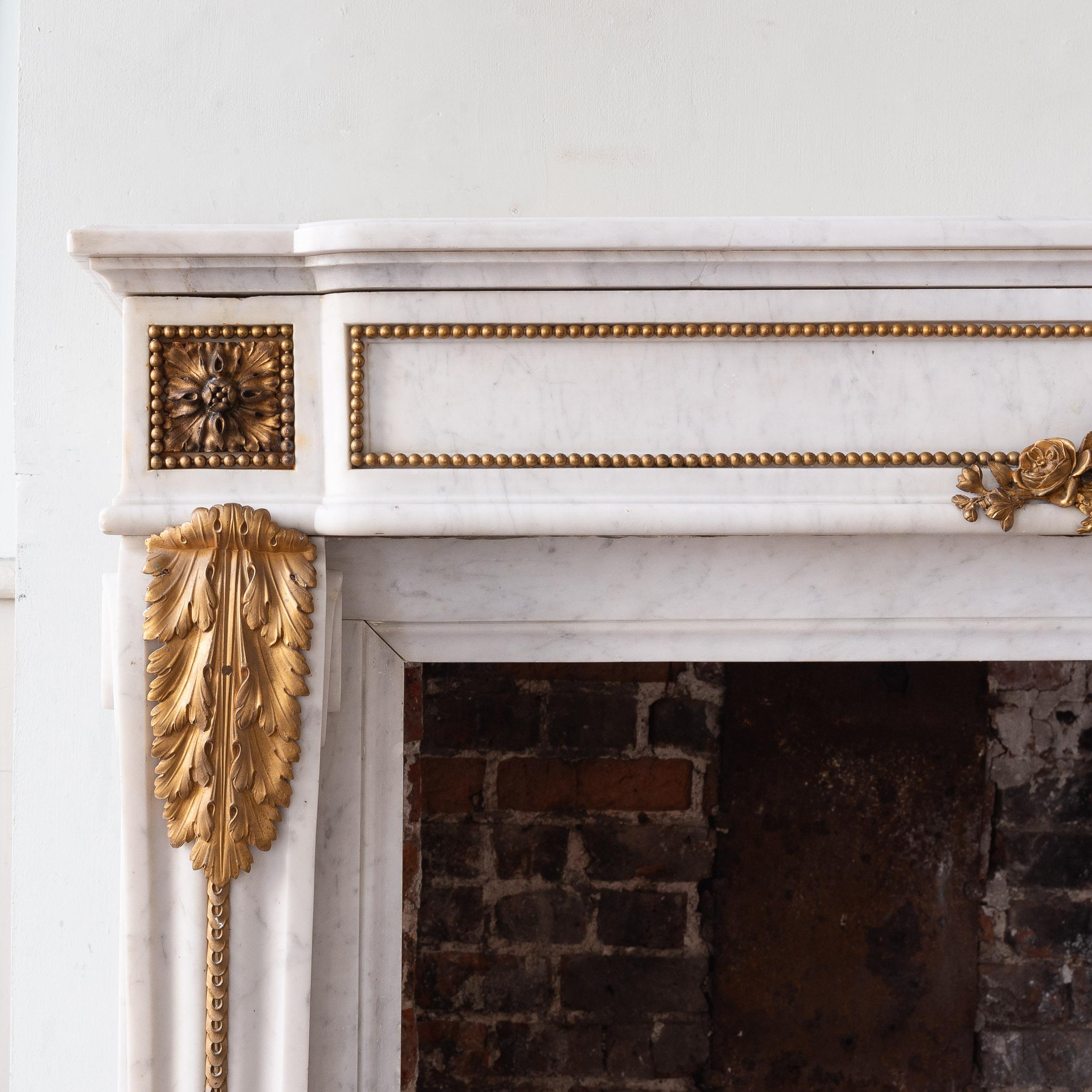A Louis XVI style Carrara marble and ormolu mounted fireplace, the shaped and moulded shelf above frieze centred by fluted panel adorned with trails of foliage and ribbons, the paterae endblocks with beaded border raised on acanthus clasped scrolled