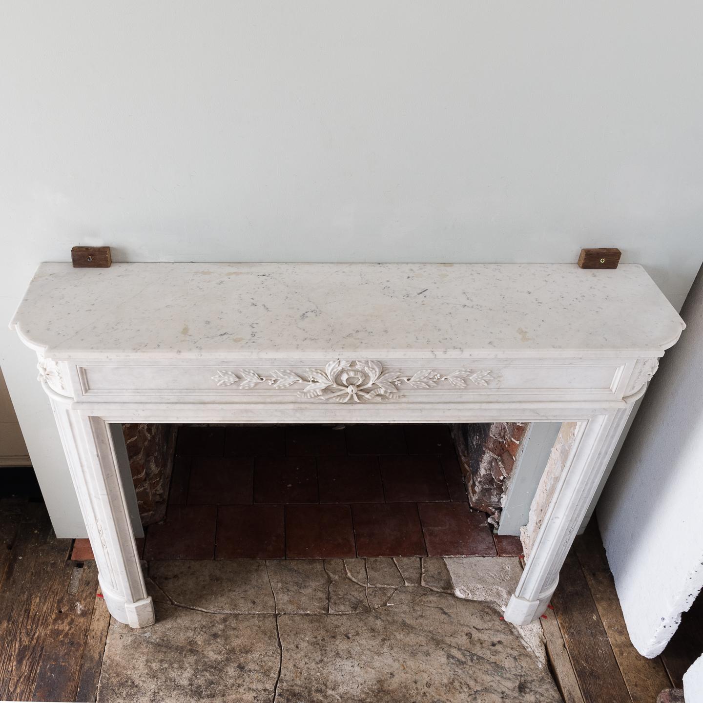 A Louis XVI style Carrara marble chimneypiece, 19th century, the moulded shaped shelf above panelled frieze centred by laurel wreath, the stop-fluted quarter-column jambs headed by square paterae corner blocks, on shaped foot blocks.
  