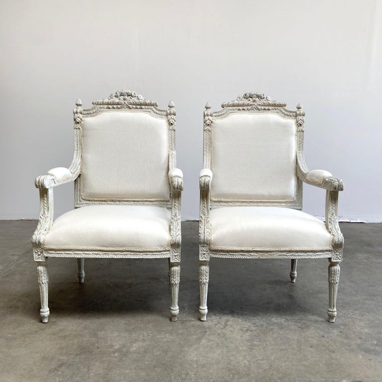 Vintage pair of painted and upholstered Louis XVI style chairs carved roses, flowers and scrolls across the top back, with a sleek fluted Louis XVI style leg. Painted in a layer of Gustavian gray, with antique white, and original gilt peeking