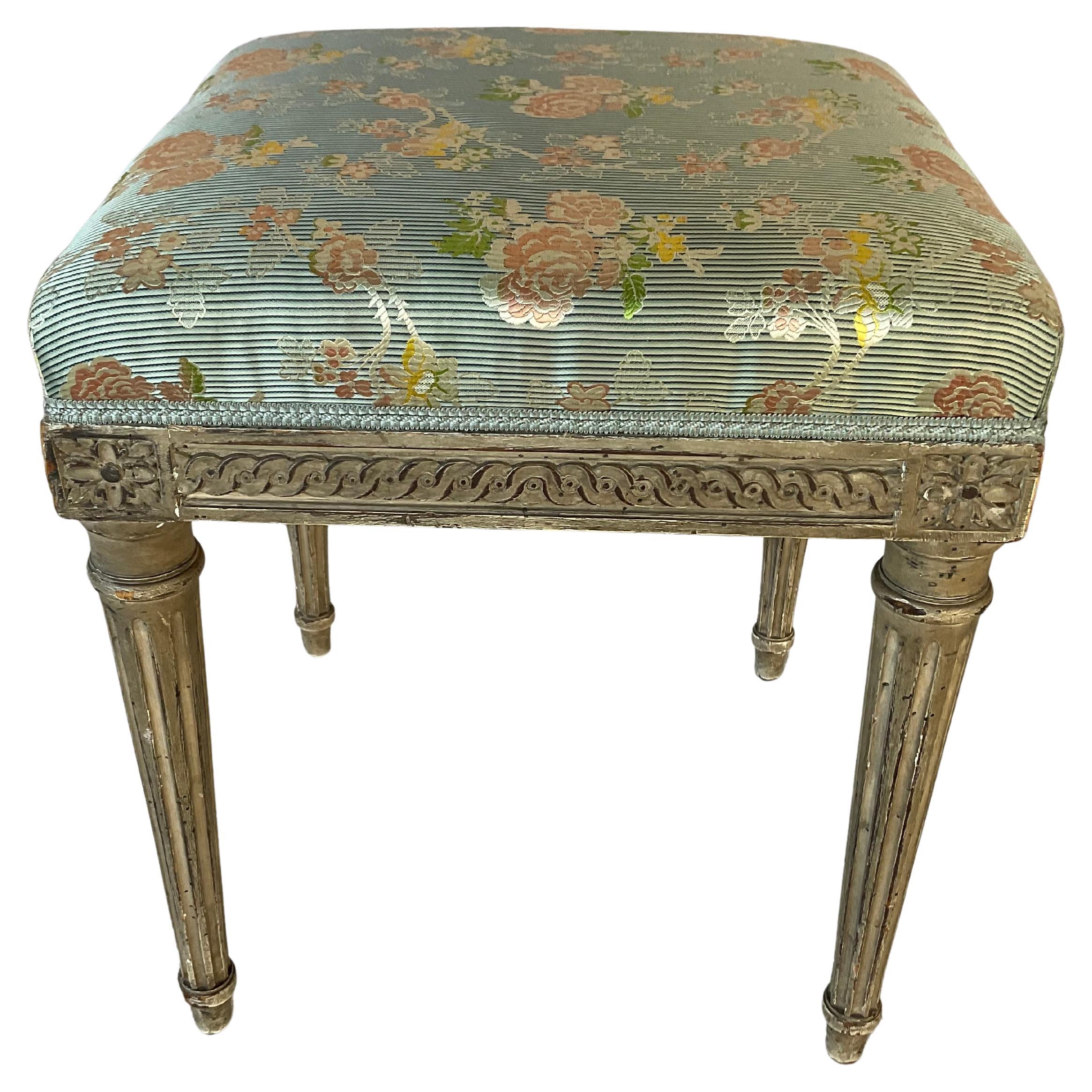 Encantador Reposapiés estilo Luis XVI tallado y pintado con tapicería de seda floral.  El marco rectangular está finamente tallado con detalles de cuentas y estrías. El taburete descansa sobre patas clásicas, cónicas y estriadas. La tapicería