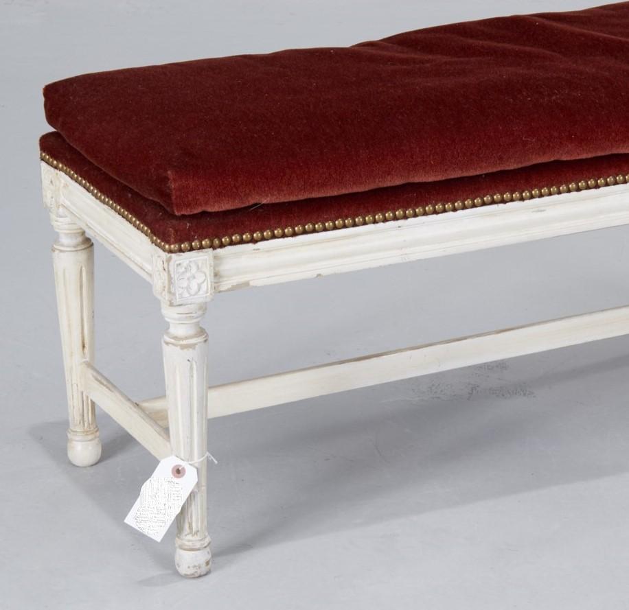 A 20th c., Louis XVI style white painted wood bench with down filled burgundy mohair seat and nail head trim. The bench is unmarked. The paint has been deliberately distressed. The legs have carved detail in both the upright column and in the upper