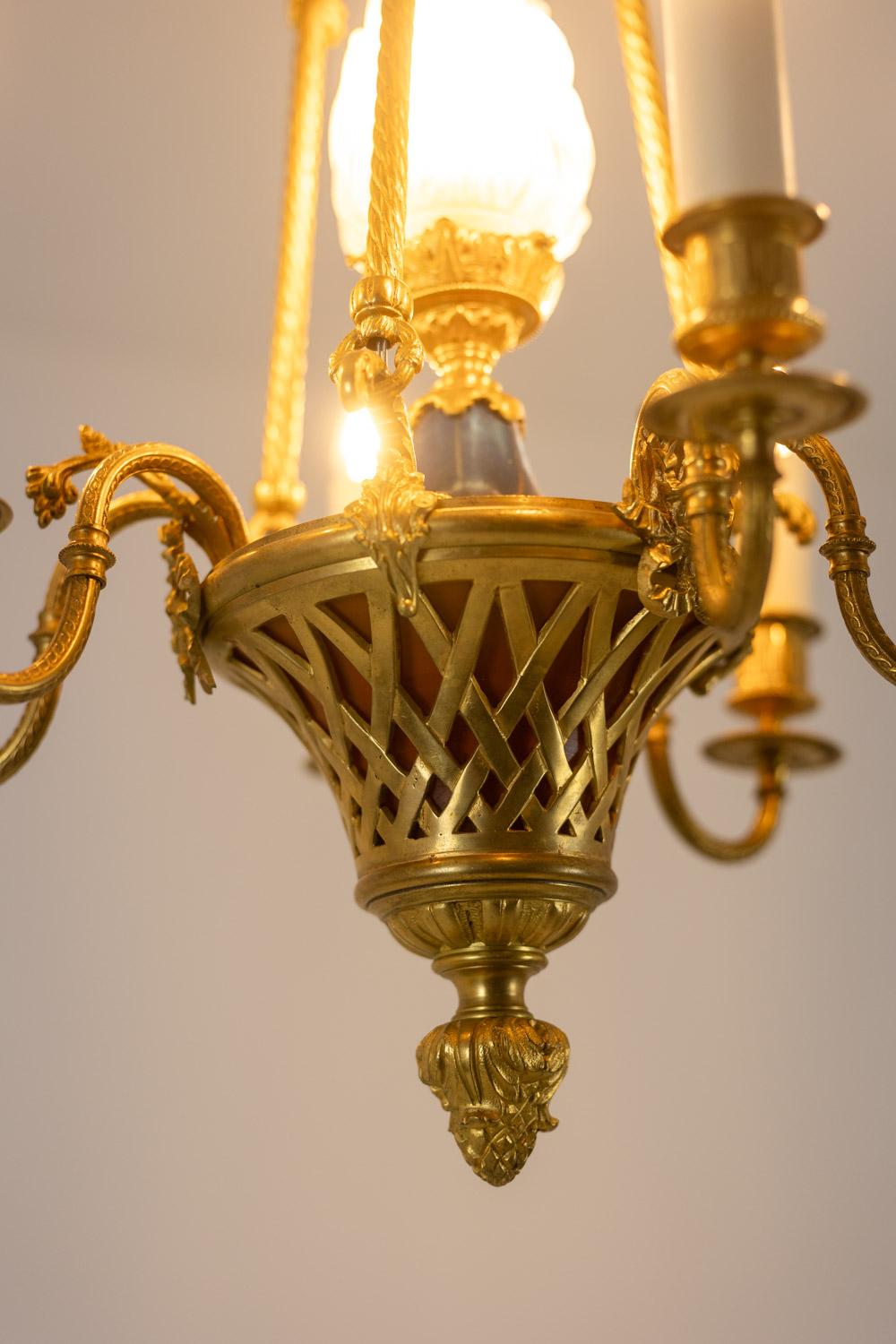 Lustre de style Louis XVI en bronze doré à six bras de lumière, groupés deux par deux par un ruban, avec leurs bobèches aux extrémités rugueuses se terminant par de fines frises régulières, qui reposent sur une coupe centrale en pâte de verre