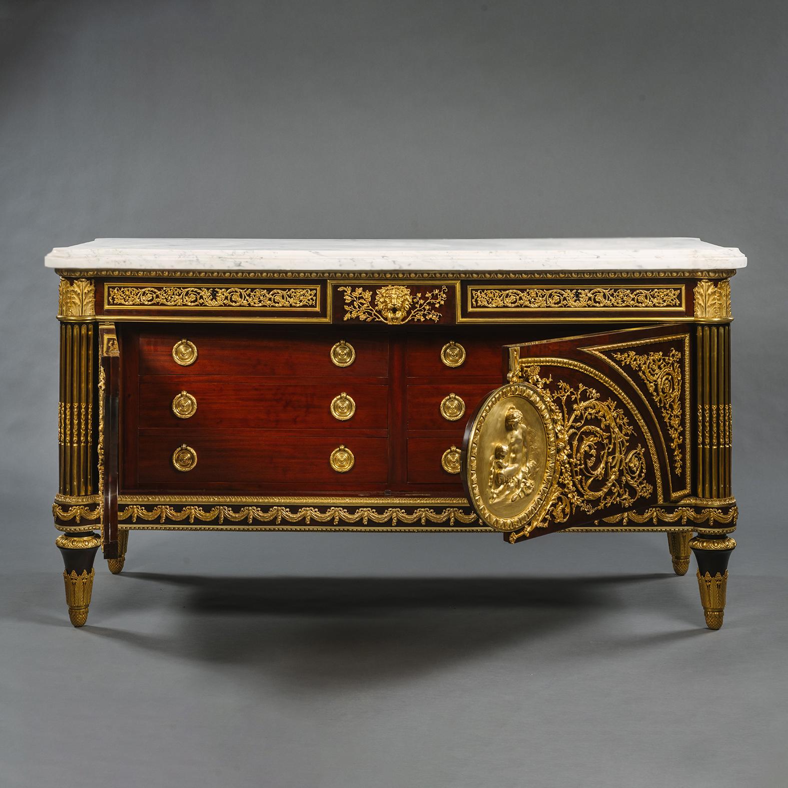 A Louis XVI Style Gilt-Bronze Mounted Mahogany Commode, After The Model By Joseph Stöckel and Guillaume Benneman. 

Decorated overall with berried scrolling foliage, the eared rectangular white marble top above egg-and-dart moulding and three frieze