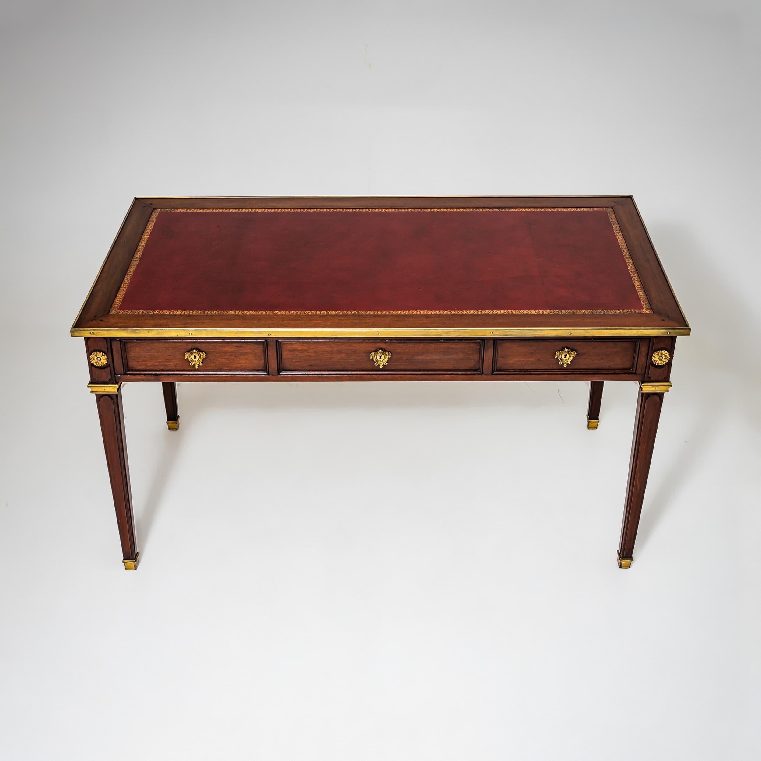 Louis XVI style desk, crafted around 1900. The tabletop features burgundy leather with a surrounding brass trim. Three shallow drawers with refined brass fittings offer practical storage. Made of mahogany, the desk stands on tall, tapering square