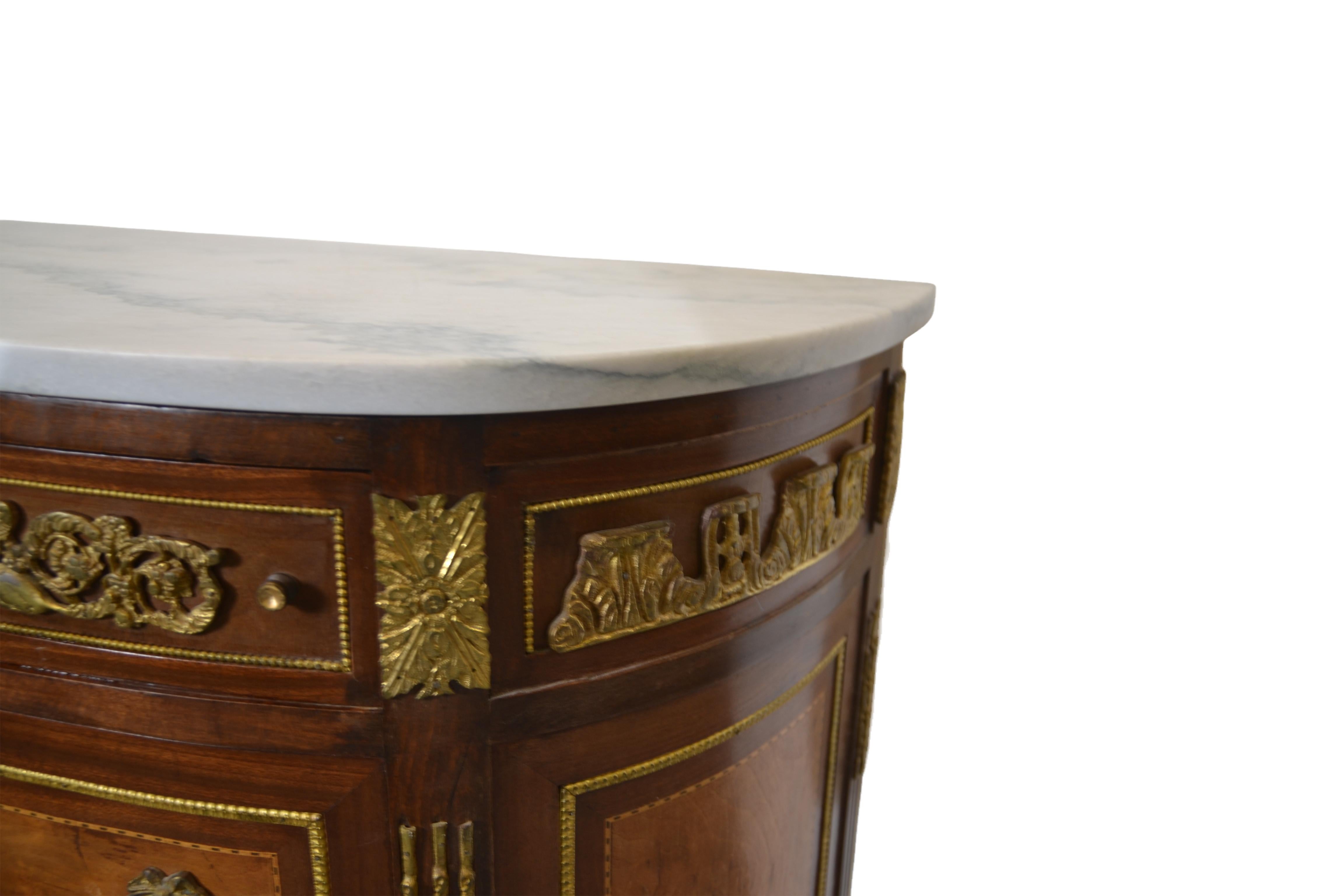 Demilune style marble top commode has dark and light wood colors. It has one small and two large drawers and detailed brass accents on the sides and the front.