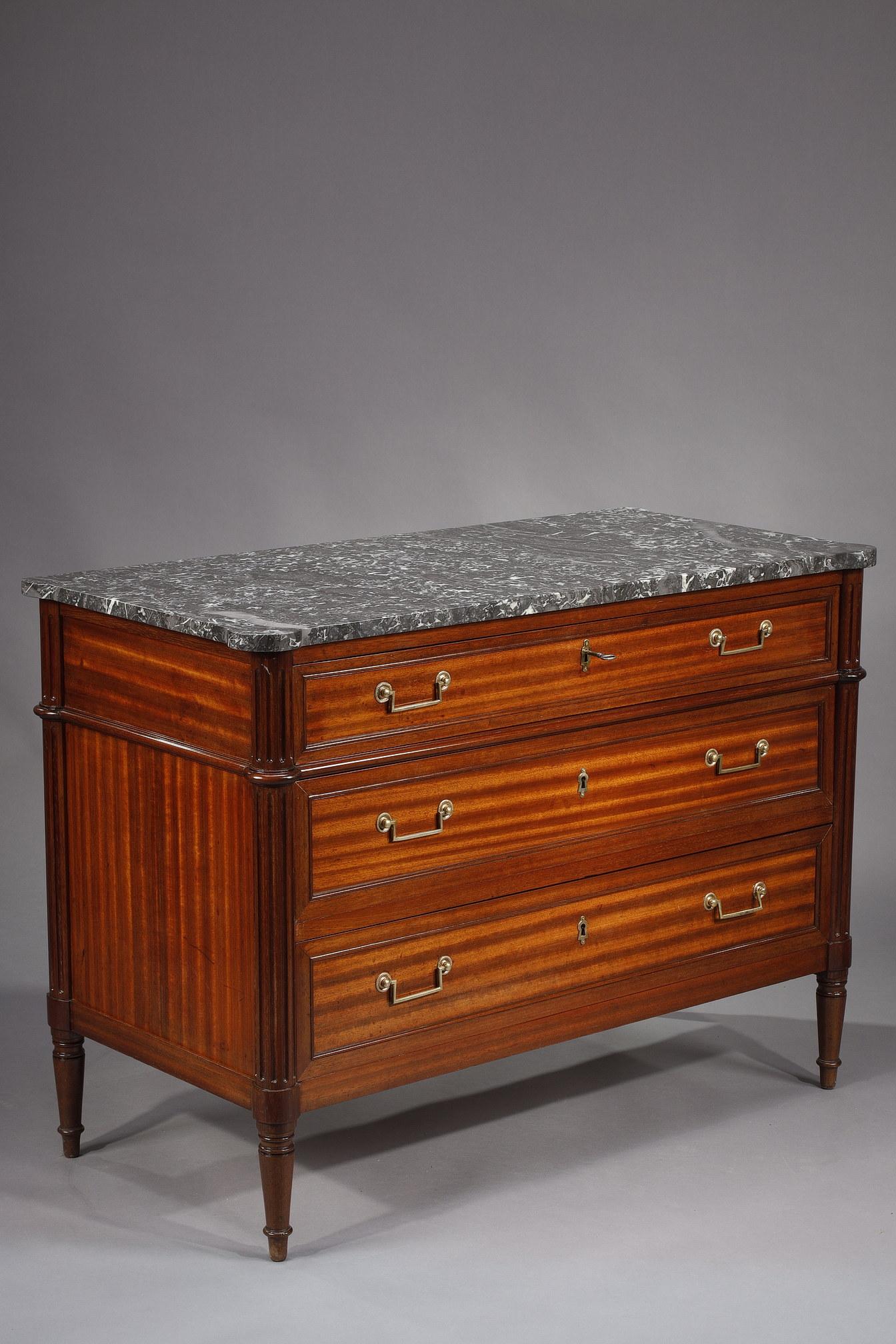 Louis XVI style chest of drawers in mahogany and flamed mahogany veneer. This piece of furniture is composed of three drawers with brass handles and fluted posts. The top is in grey Saint-Anne marble. The unit rests on turned and tapered legs.