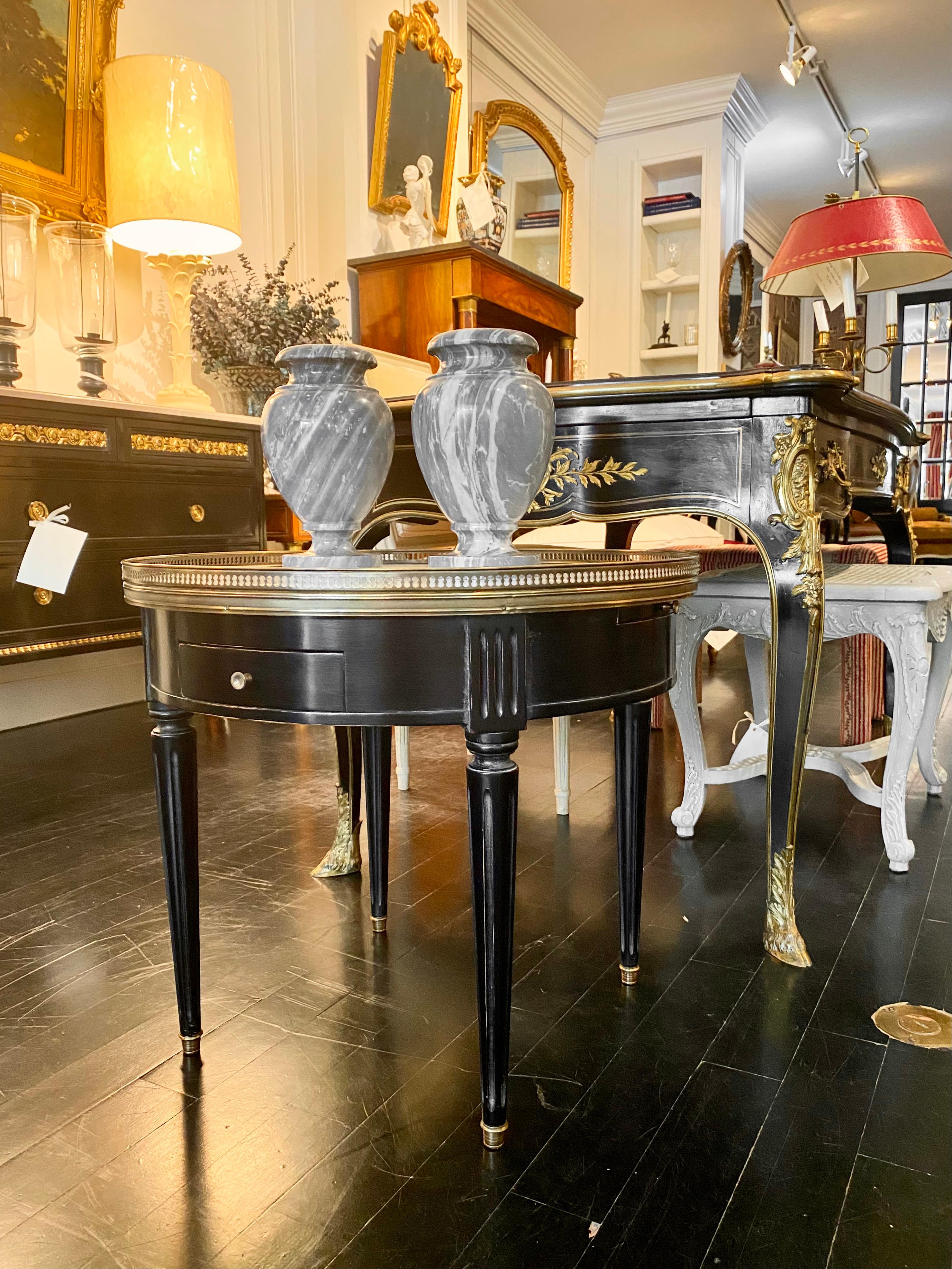 Louis XVI style ebonized marble-top bouillotte table.
Elegant French round bouillotte table. Classic Louis XVI style. Two drawers and two pull-out tirelles slides. White carrara marble top is surrounded by a pierced bronze gallery. The table sits