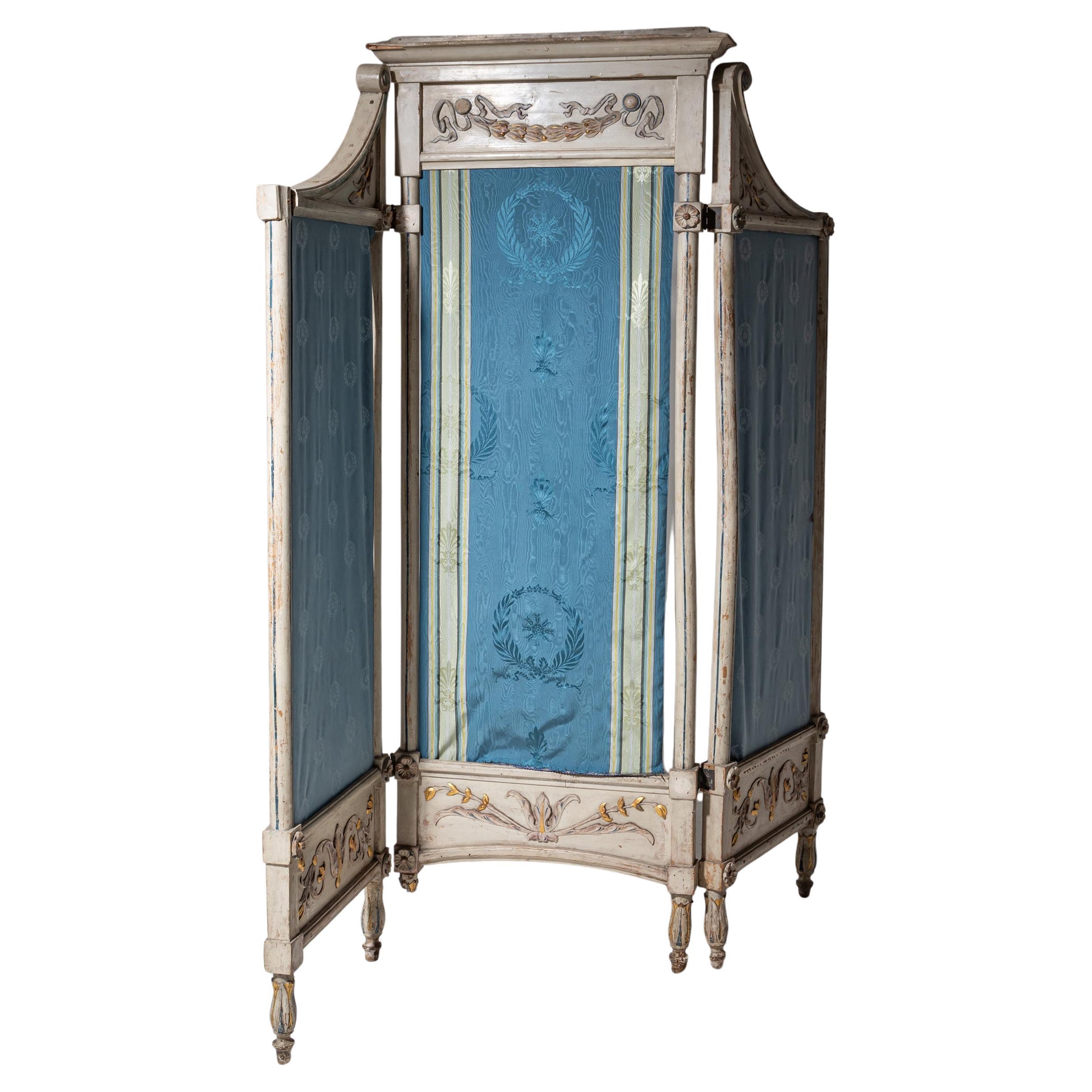 Paravento pieghevole a tre pannelli con una tenda di seta blu moiré con ornamenti di alloro. La cornice dipinta di grigio è decorata con rosette e motivi di foglie. La superficie mostra i segni dell'età e dell'uso.