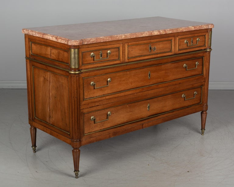A Louis XVI style French commode, or chest of drawers, made of solid walnut with brass trim and original pink marble top. Five dovetailed drawers with original pulls and working locks with one key. Fluted columns with rounded front corners ending in