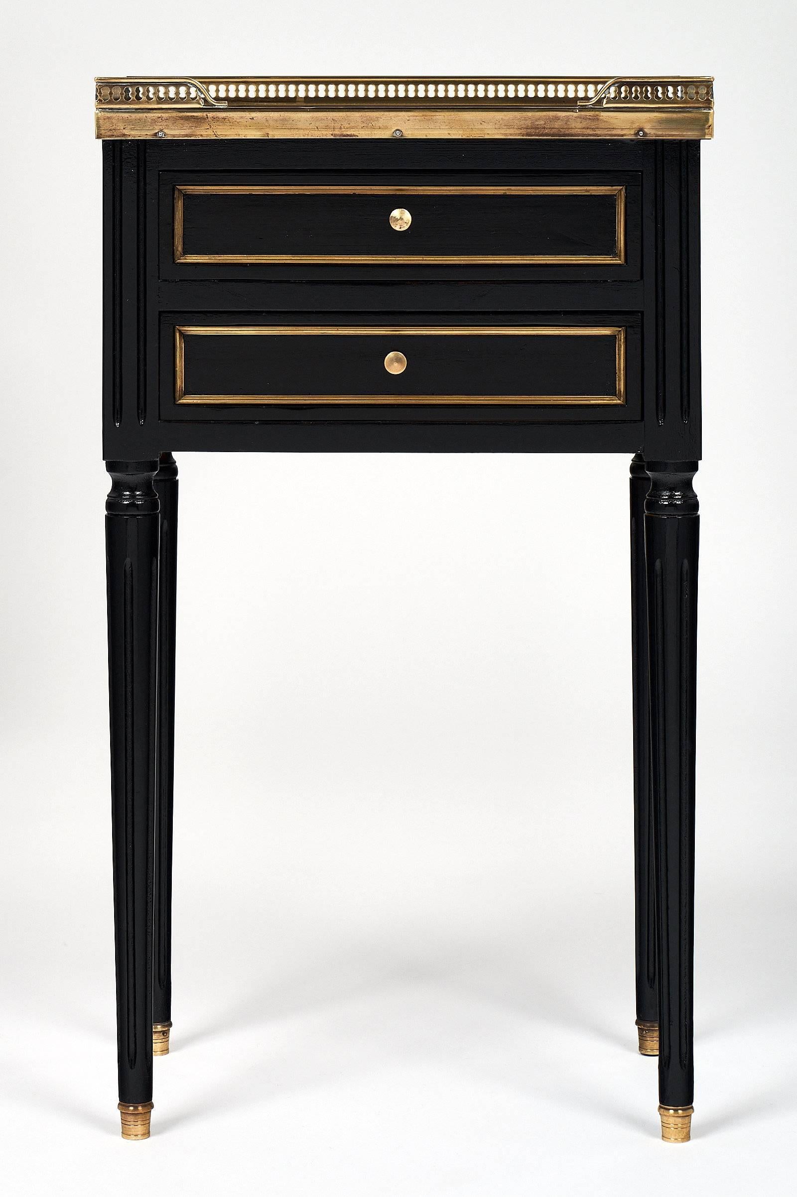 A pair of Louis XVI side tables made of ebonized mahogany that has been finished with a lustrous French polish. Each table is topped with Carrara marble, and features a gilt brass gallery, two dovetailed drawers; and fluted legs with brass feet. An