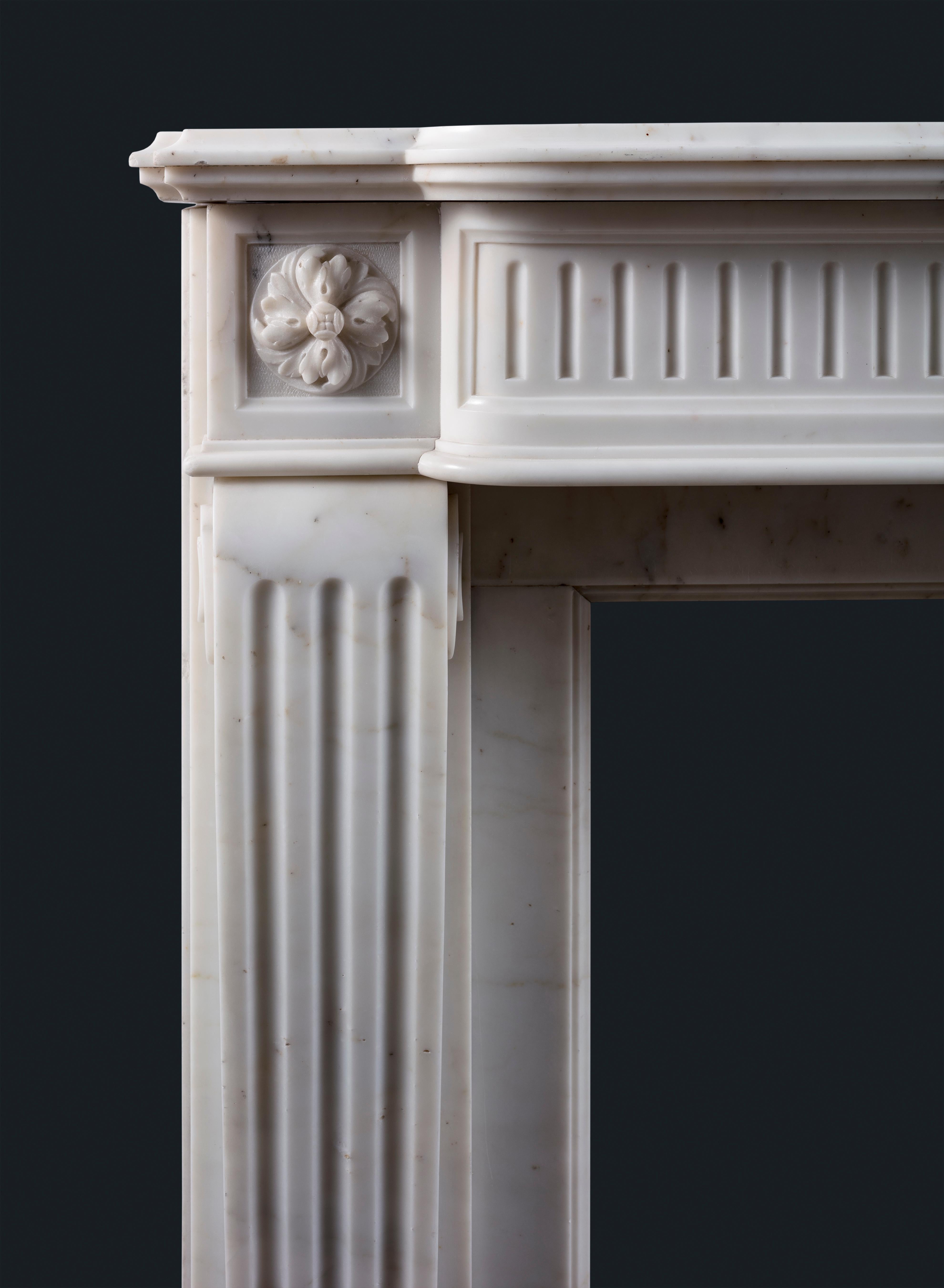 An antique Louis XVI-style fireplace surround, the generously shaped and moulded shelf above bow-front fluted frieze with foliate carved central plaque and foliate paterae to each flank, all raised on stop-fluted scroll jambs and block feet, in