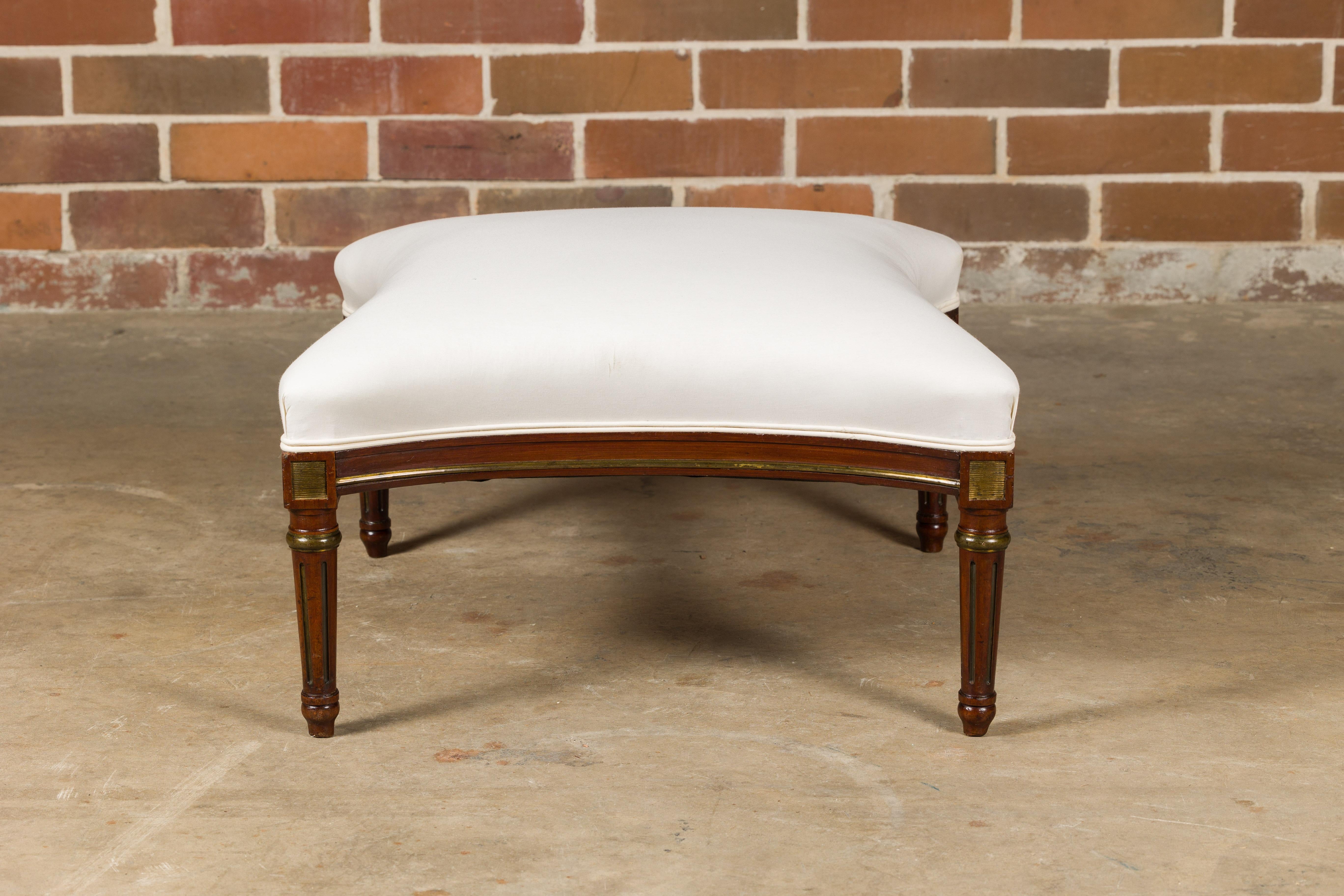 A French Louis XVI style wooden ottoman from circa 1900 with fluted tapering legs, gilded accents and muslin double welt upholstery. Immerse yourself in the elegance of French design with this Louis XVI style wooden ottoman from circa 1900, a piece