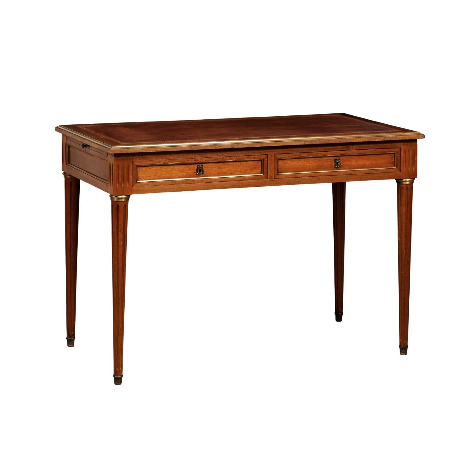 A French Louis XVI style walnut desk with leather top, pull-outs on the sides, two drawers, carved fluted legs and brass trim. Steeped in timeless French elegance, this Louis XVI style walnut desk, hailing from the 20th century, is a testament to