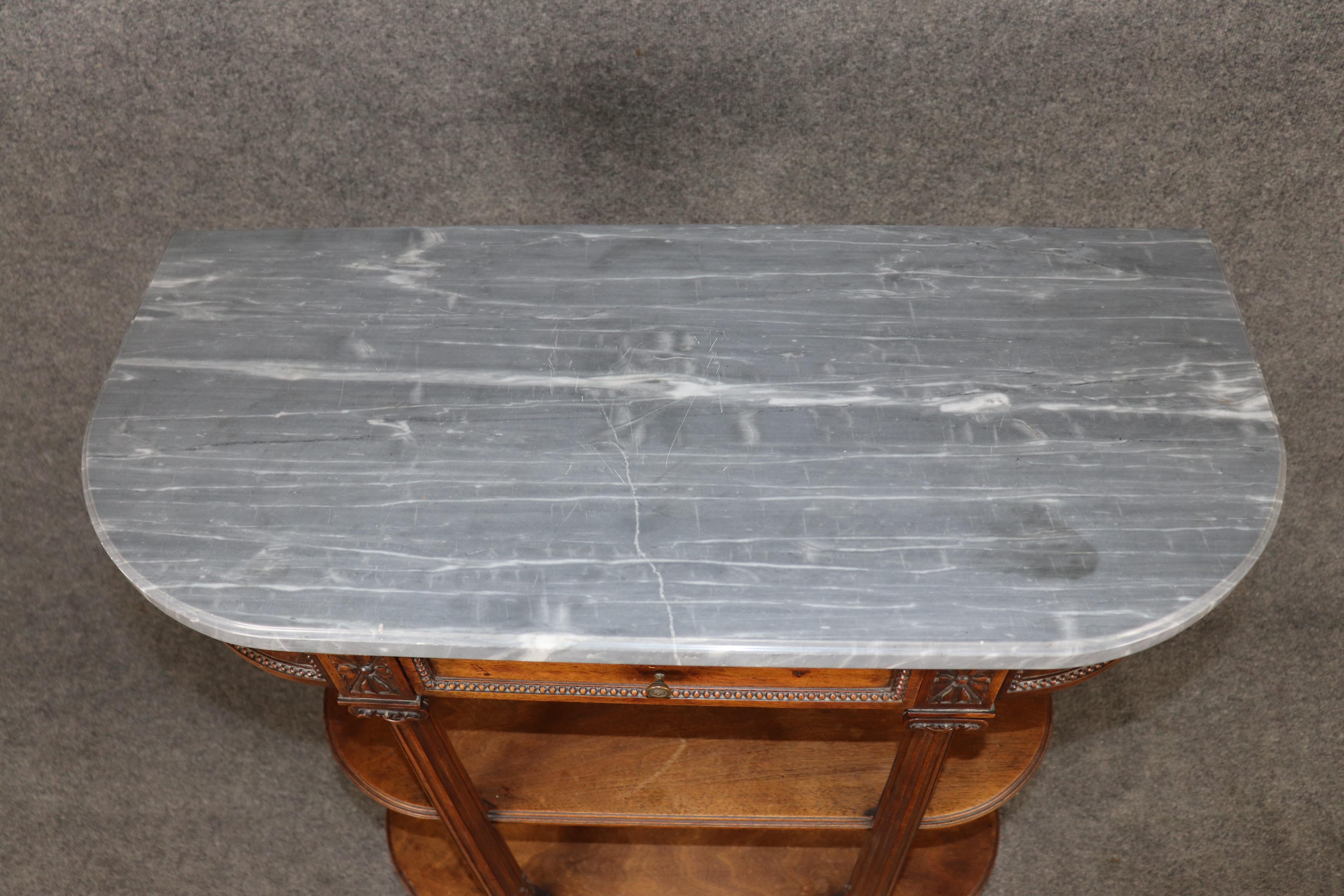 
This elegant French three-tier console table from the 1910s combines timeless neoclassical style with functional design. The piece features a polished marble top in soft gray tones, gracefully contrasted by a warm walnut frame. Its serpentine front