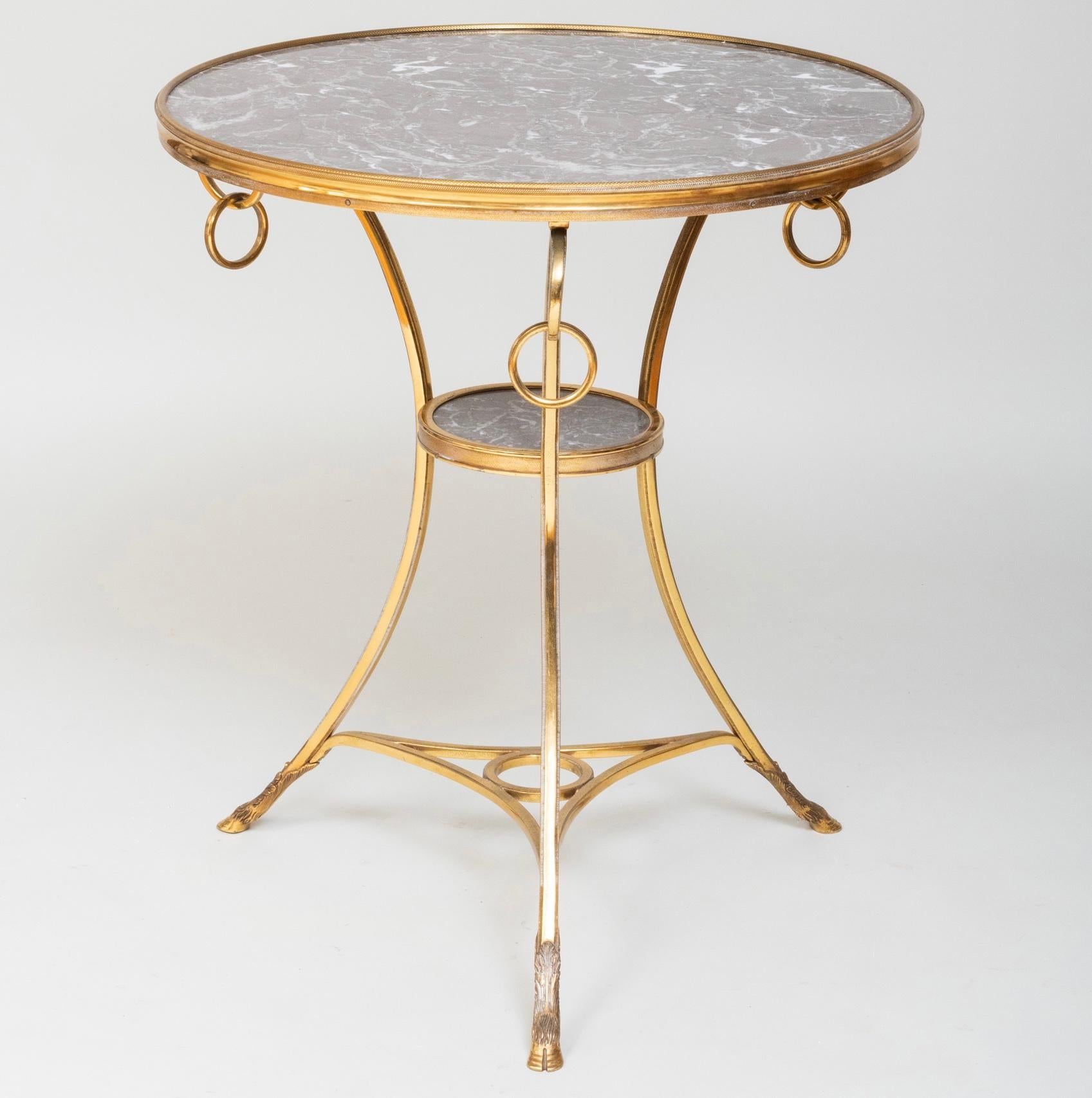 Elegante table d'appoint ronde avec des anneaux décoratifs suspendus au tablier, et des pieds fourchus. Détails et qualité raffinés.