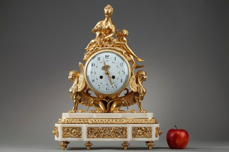Gilt bronze clock in the Louis XVI style representing a nymph and two cherubs. The nymph, undressed and resting on a cushion, is nursing one of the cherubs while the second one calls her. In the center, the round enamelled dial is decorated with