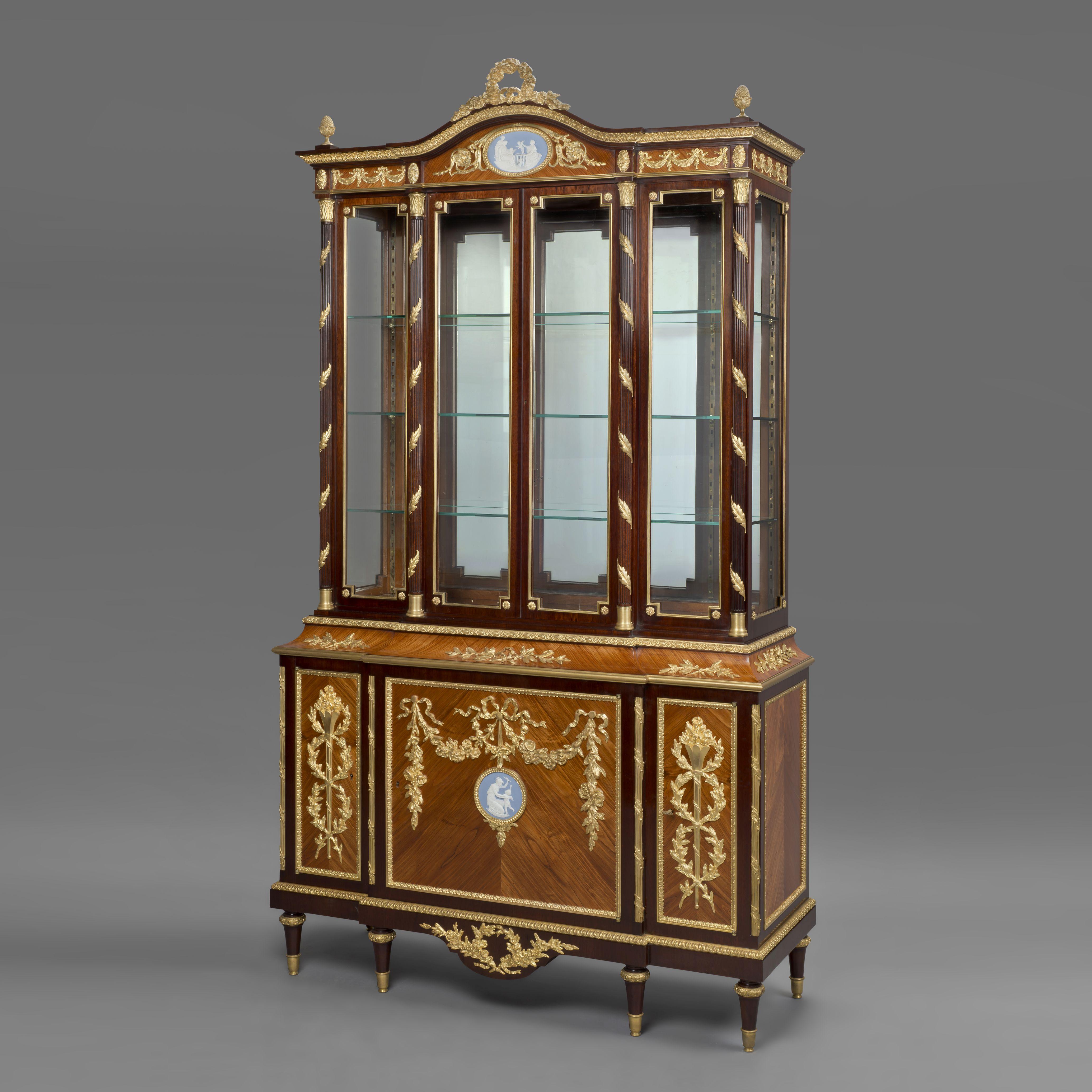 A Fine Louis XVI Style Gilt-Bronze Mounted Mahogany Display Cabinet With Wedgwood Porcelain Plaques.

The upper section has a shaped cornice with acorn finials and a foliate wreath above a frieze centred by a large oval Wedgwood porcelain plaque. 