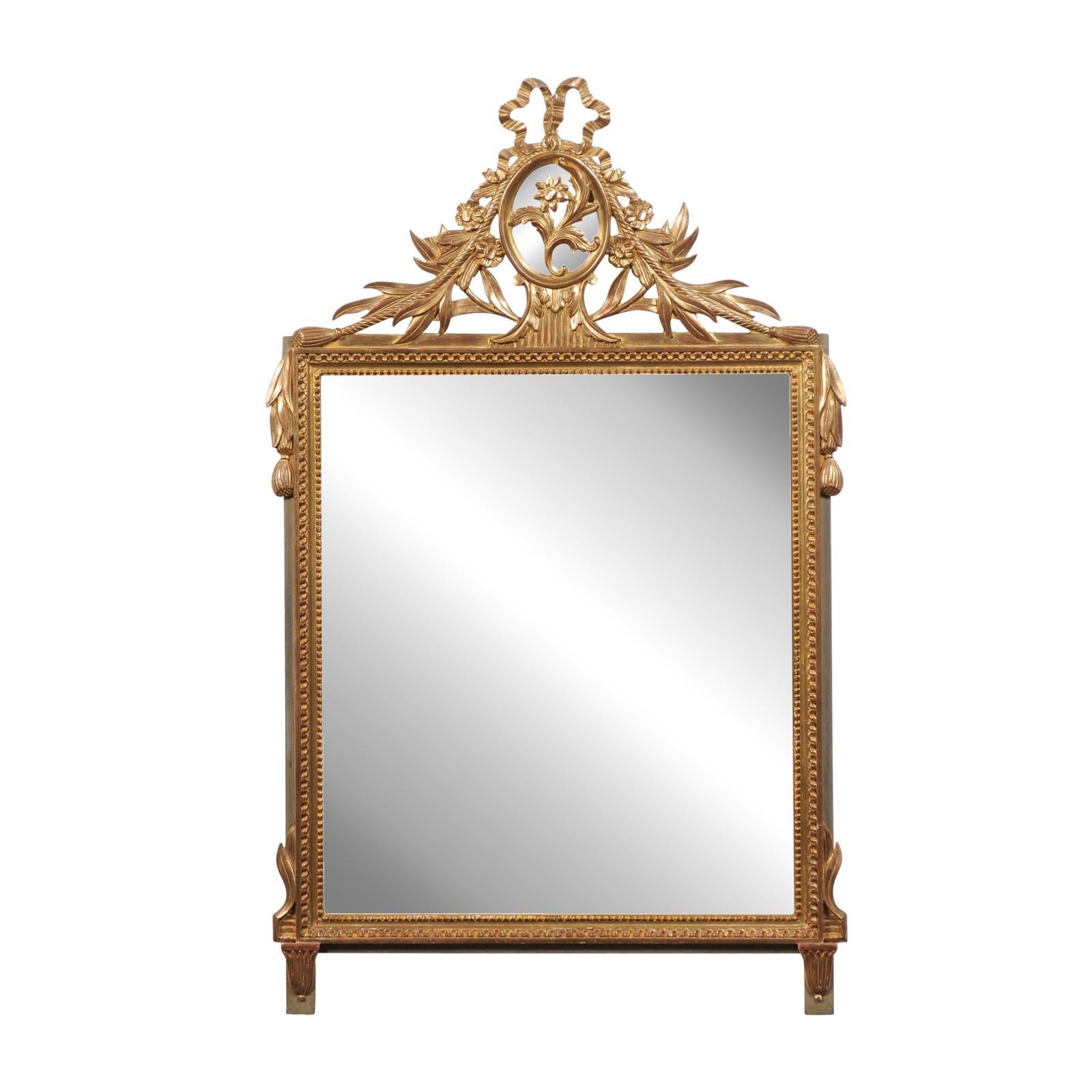 A French Louis XVI style gilded wood mirror from the 20th century with ribbon-tied floral medallion, petite carved beads and tassels. Exuding the grace and grandeur of French craftsmanship, this Louis XVI style gilded wood mirror from the 20th