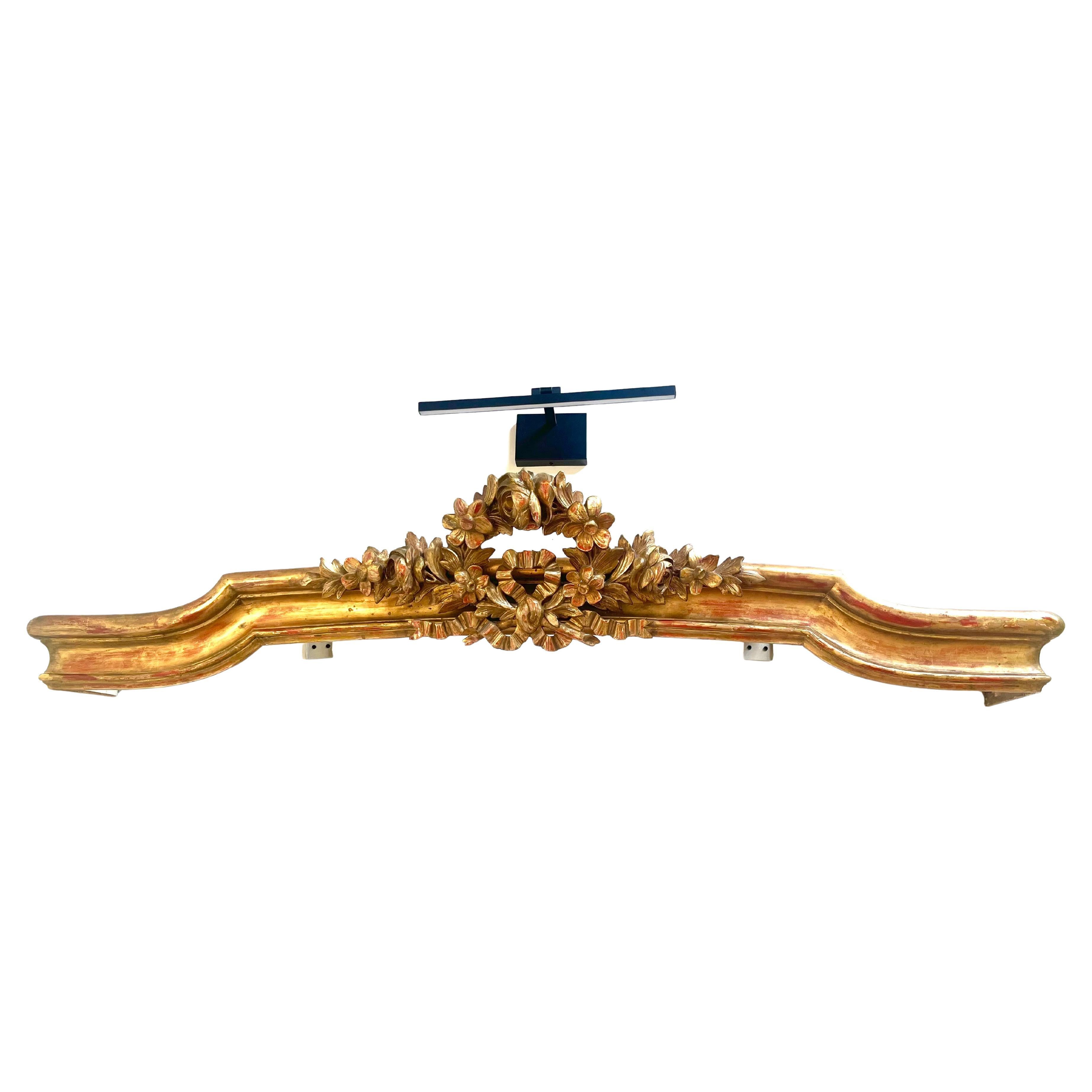 Ein vergoldetes architektonisches Element, wahrscheinlich ein Overdoor (Fenster), jetzt als Kranz oder Betthimmel montiert. Ein zentrales Blumenbouquet vor einem gestuften und geformten Gesims, die geschwungenen Enden mit einem Eisenflansch oder