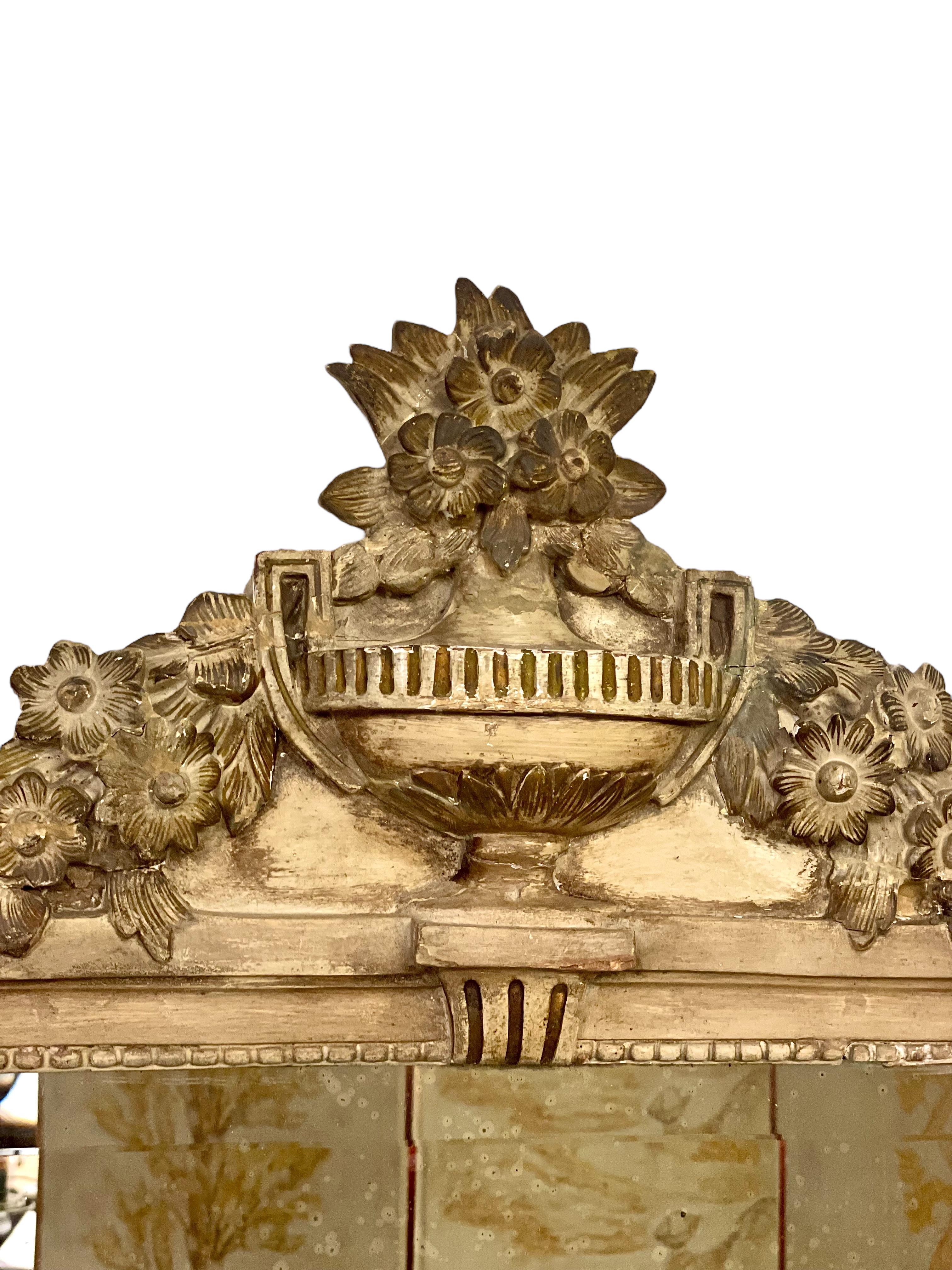Ce miroir mural très décoratif de style Louis XVI est sculpté à la main avec des motifs étonnamment détaillés, et magnifiquement fini dans une laque grise sourde. Il est couronné d'un fronton orné qui prend la forme d'un large vase, d'où partent en