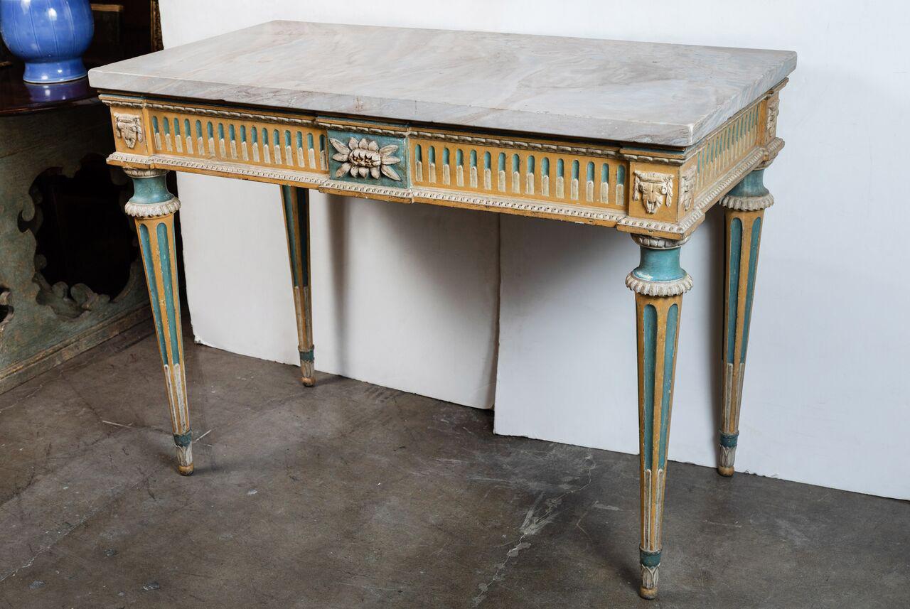 Wonderful pair of ochre and seafoam painted Louis XVI style consoles in old wood. Each with relief carved aprons featuring foliate medallions above fluted, tapered legs. Both surmounted by thick slabs of crème and grey marble.