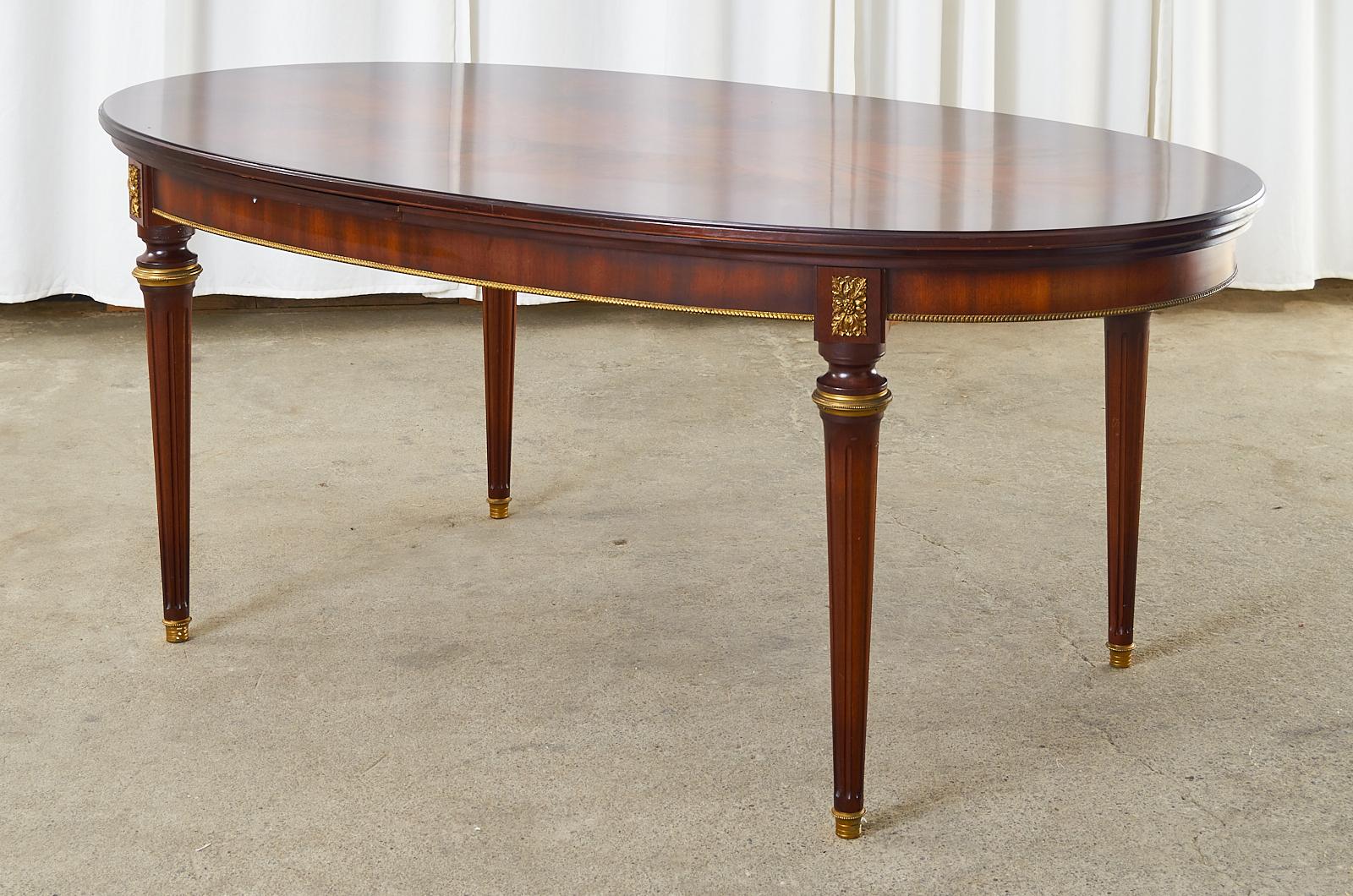 Grand French Louis XVI Style mahogany oval dining table featuring bronze ormolu mounts. Beautifully crafted with a flame match veneered top and hidden draw leaves on each side. The top measures 72 inches closed and extends to 108 inches open. Each
