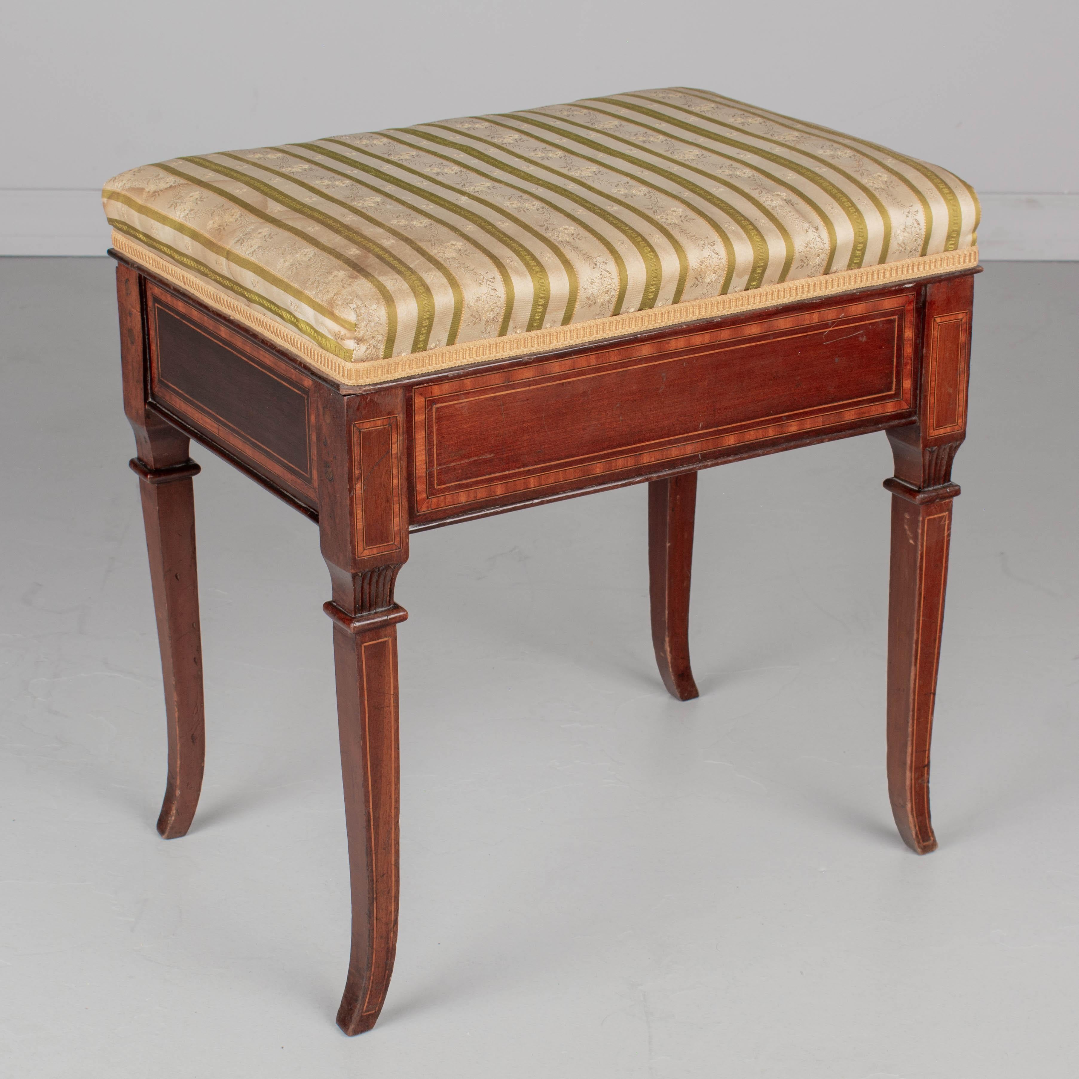 A Louis XVI style French mahogany piano bench with upholstered seat. Made of solid mahogany with marquetry inlay. Seat hinges open to reveal storage for sheet music. Original silk fabric is lightly soiled. Circa 1950s. 
18.5