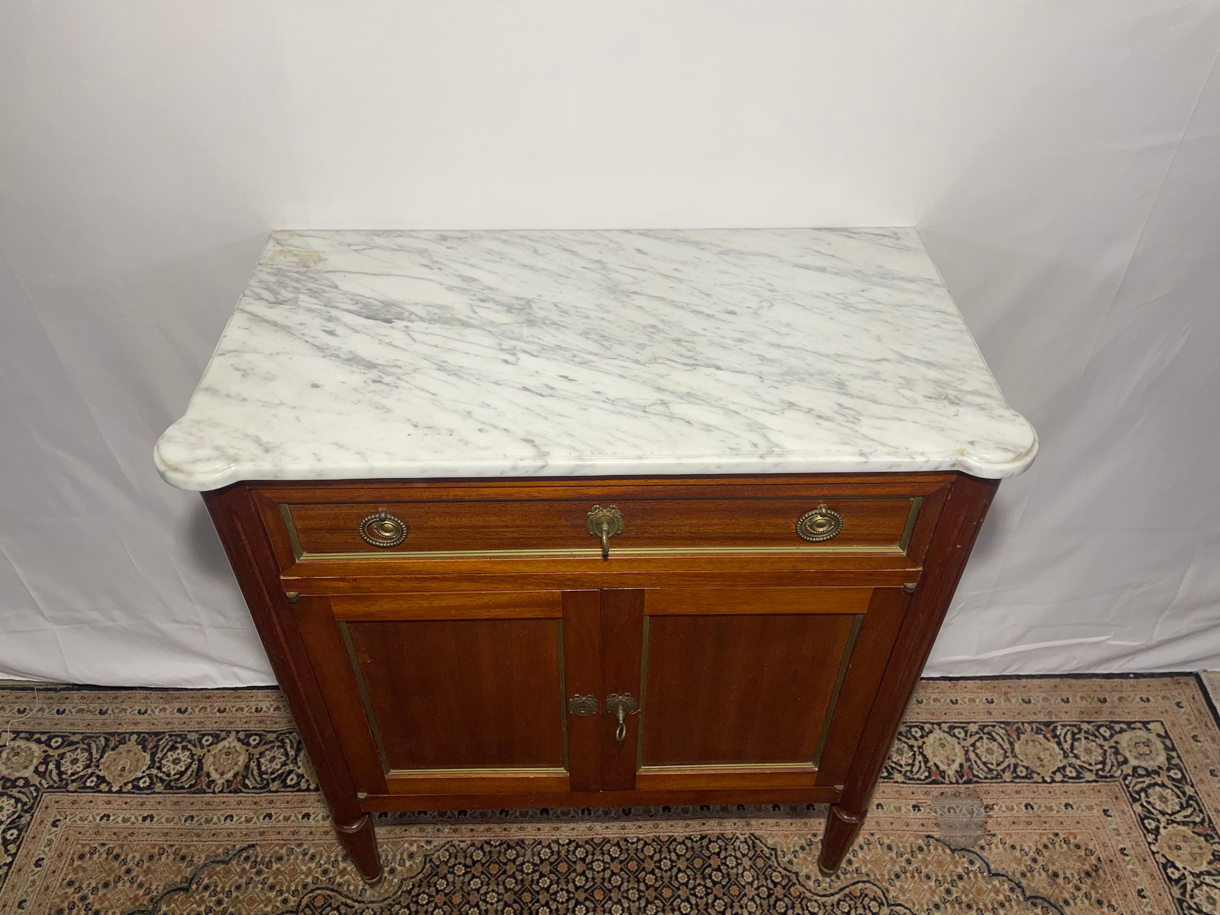 Cet élégant buffet de style Louis XVI du début du XXe siècle présente un cadre en acajou, un plateau en marbre blanc raffiné et des ferrures en laiton. Les lignes épurées, les pieds cannelés et les incrustations de laiton soulignent son design
