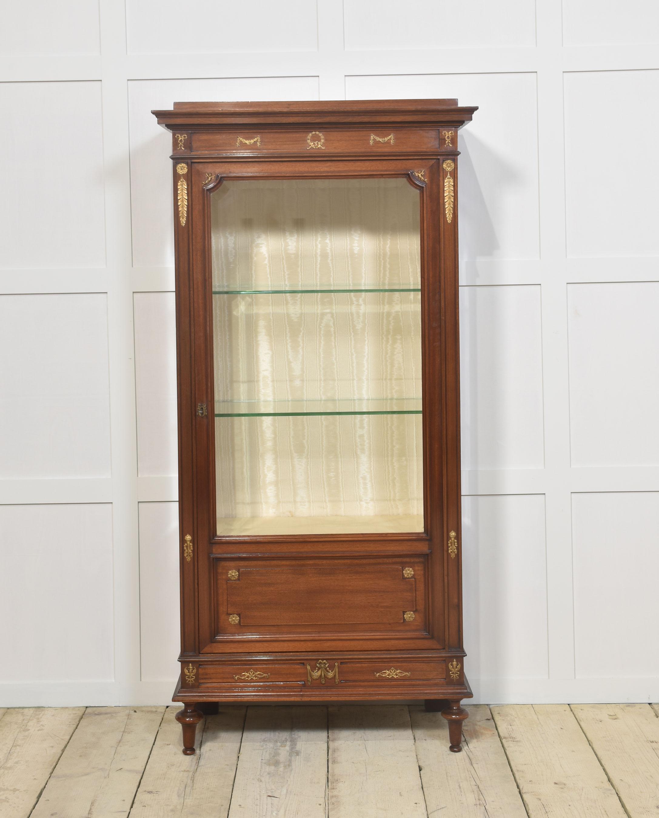 Louis XVI designed mahogany display cabinet. The projecting stepped cornice over recessed frieze decorated with gilt metal mounts, to the large bevelled glazed door opening to reveal silk upholstered interior having two glass shelves. All raised on