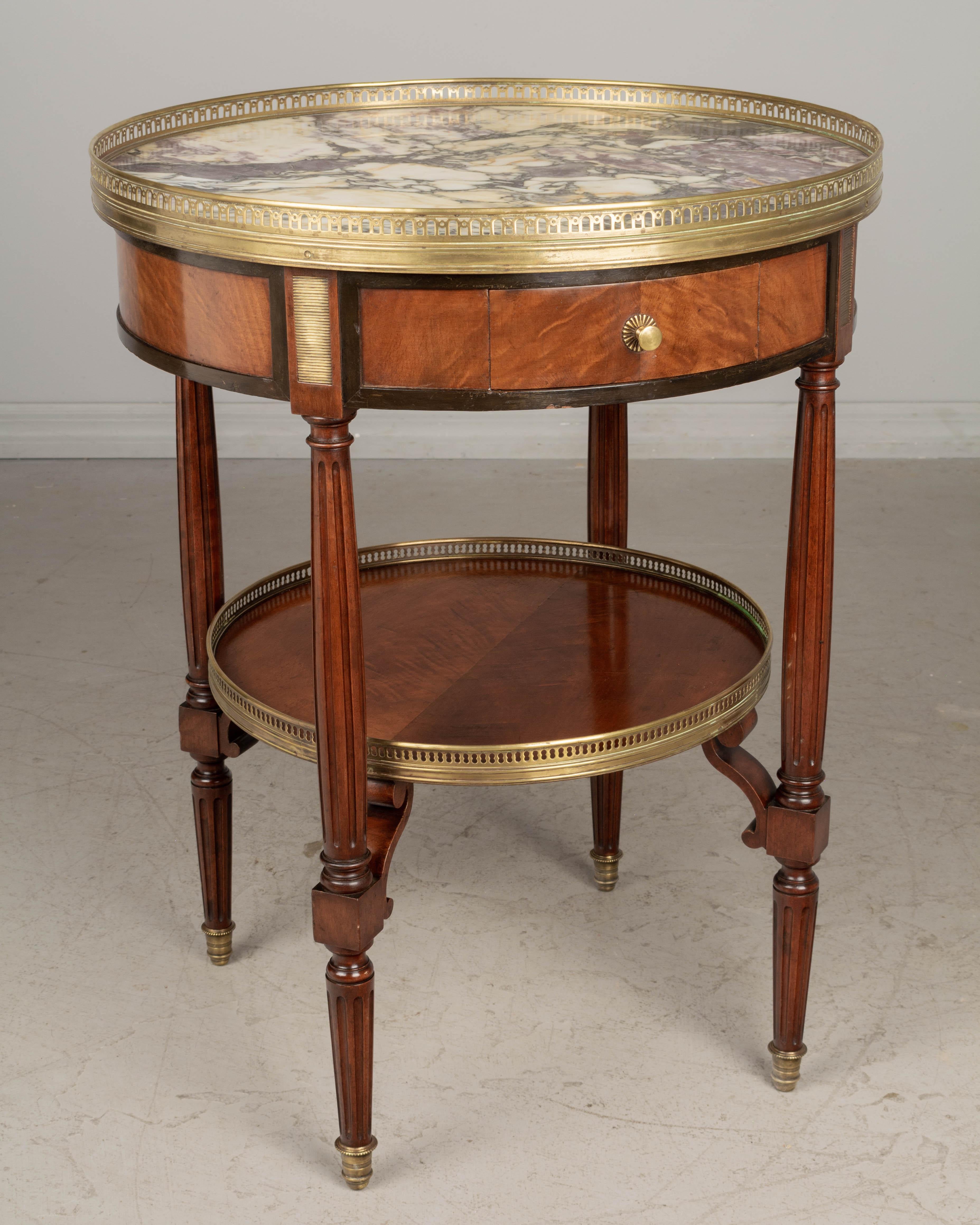 A Louis XVI style marble top bouillotte table made of solid and veneer of mahogany with a small dovetailed drawer, fluted tapered legs and lower shelf. Marble top from the Pyrenées surrounded by a brass galleries and decorative elements and sabots.
