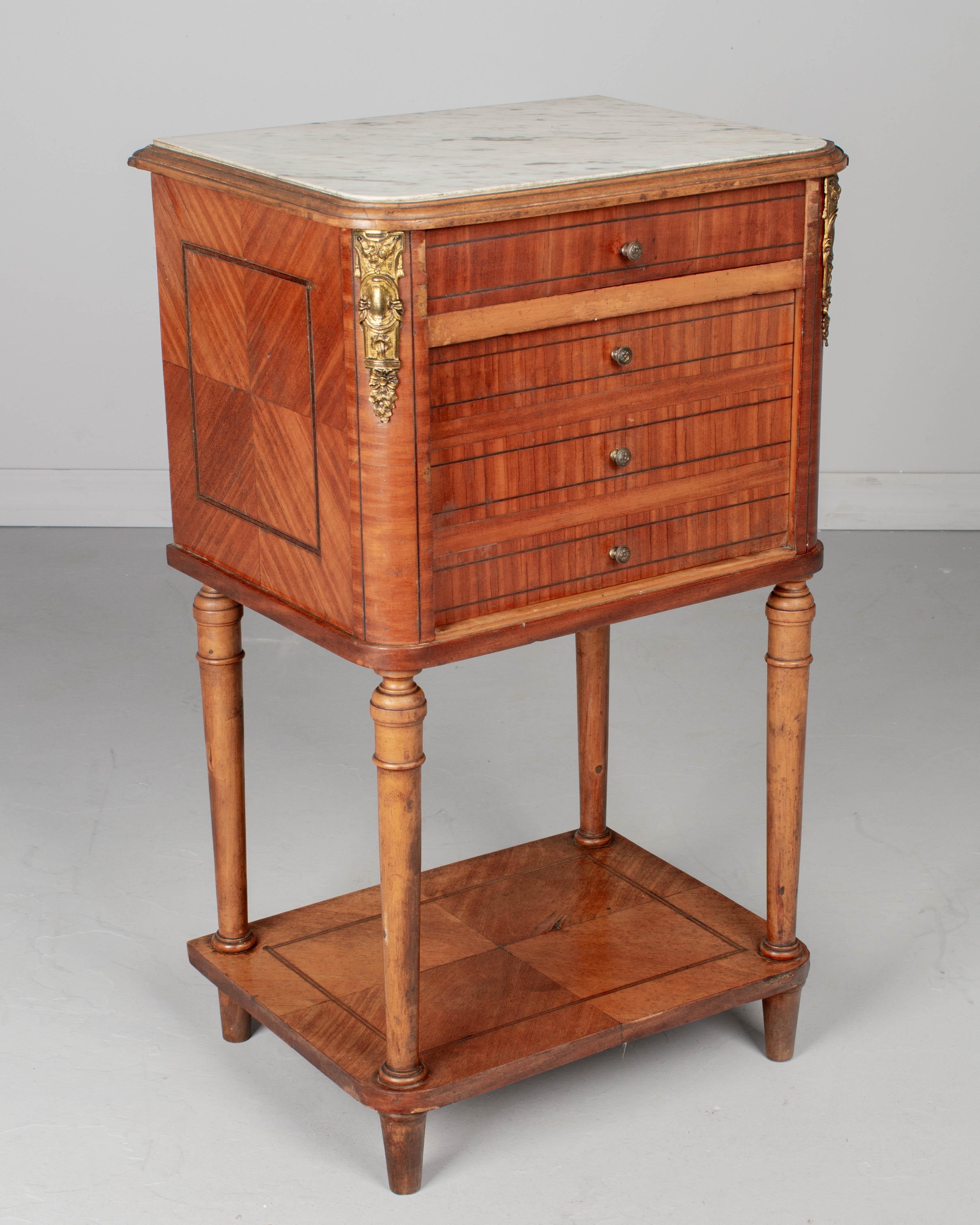 A Louis XVI style marble top marquetry nightstand, or side table, made of solid and veneer of mahogany with a small dovetailed drawer, turned tapered legs and lower shelf. Cabinet door with false drawer front opens to a marble lined interior.