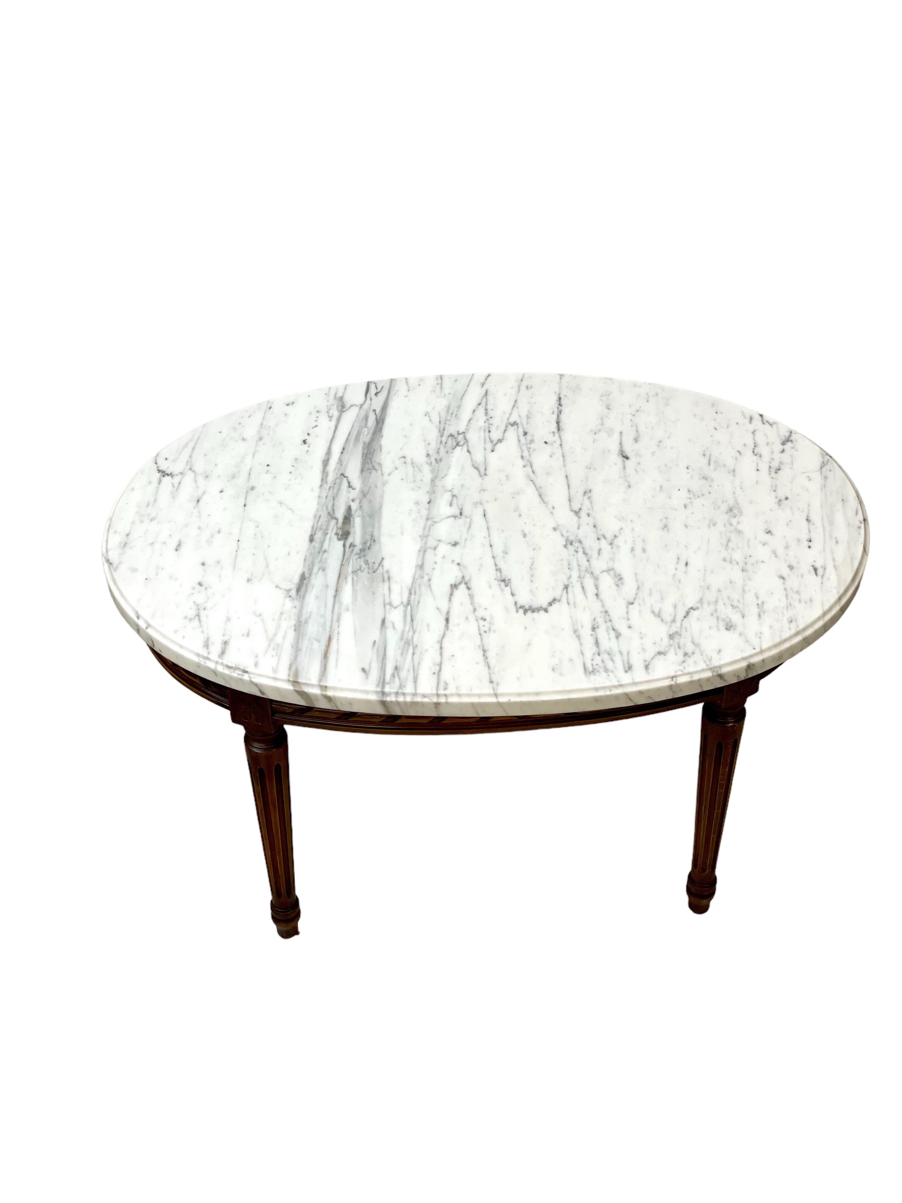 Très belle table basse ovale de style Louis XVI avec un superbe plateau en marbre blanc et gris veiné. Une frise simple mais élégante de style corde torsadée, sculptée à la main, orne le tablier en bois bruni, qui relie les quatre pieds fuselés et