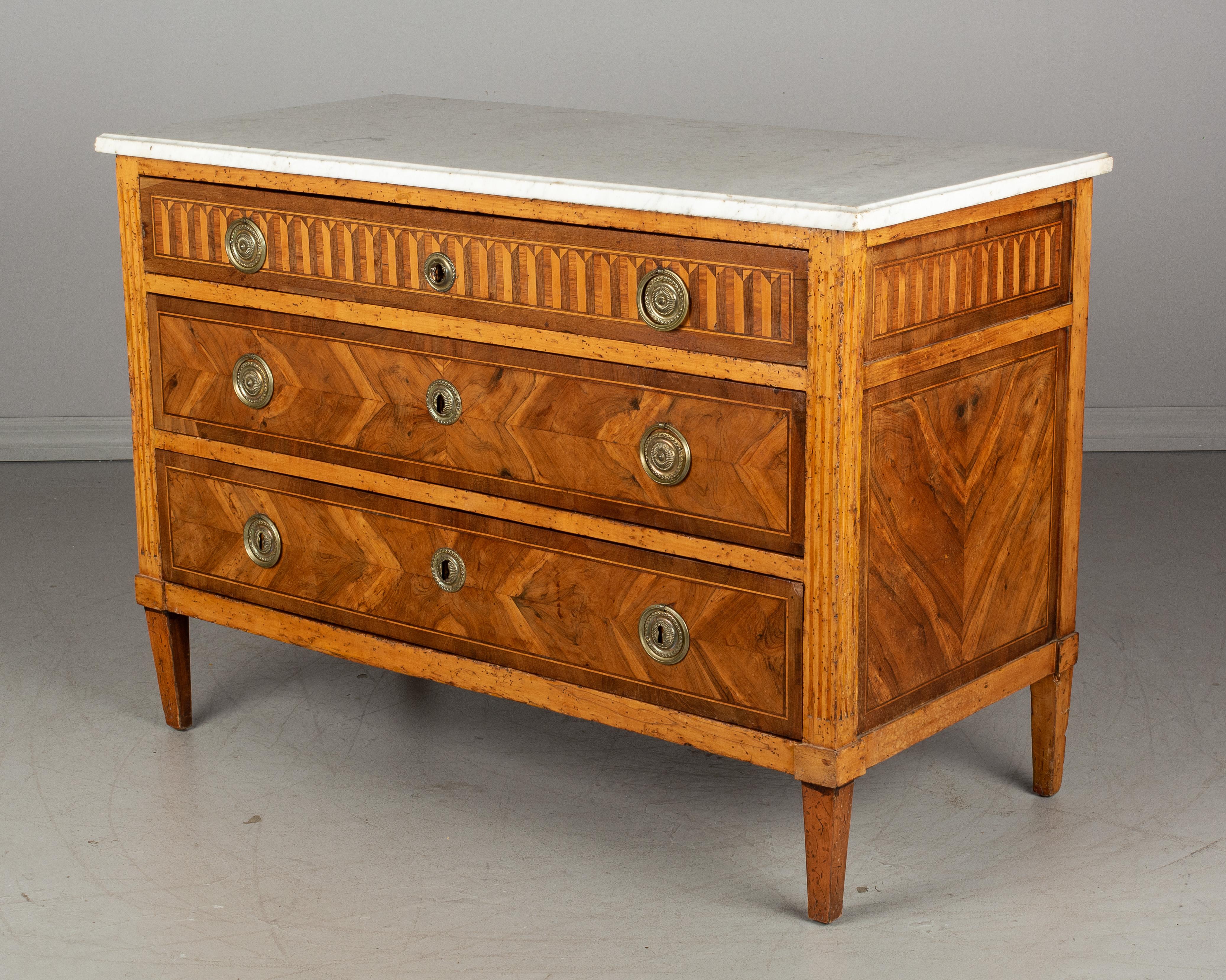 A 19th century French Louis XVI style marquetry commode with three dovetailed drawers and white marble top. Made of solid walnut with bookmatched veneer of walnut and various wood inlay. Original brass hardware. Locks are not present and there are