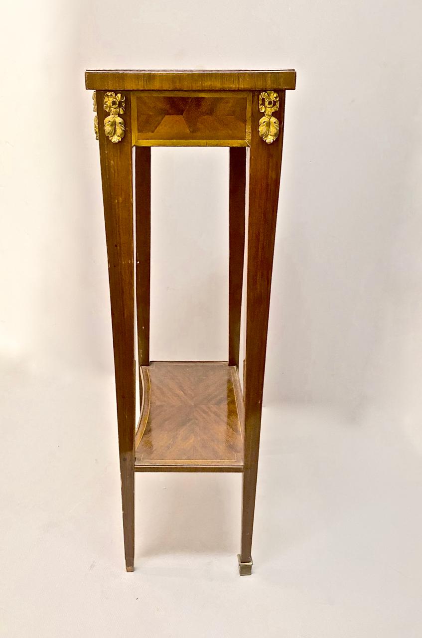 This is a chic Louis XVI-style stand that is detailed with finely executed marquetry and gilt mounts. The single drawer includes it's key. The stand dates to the mid-19th century. Two of the brass sabots are replacements.