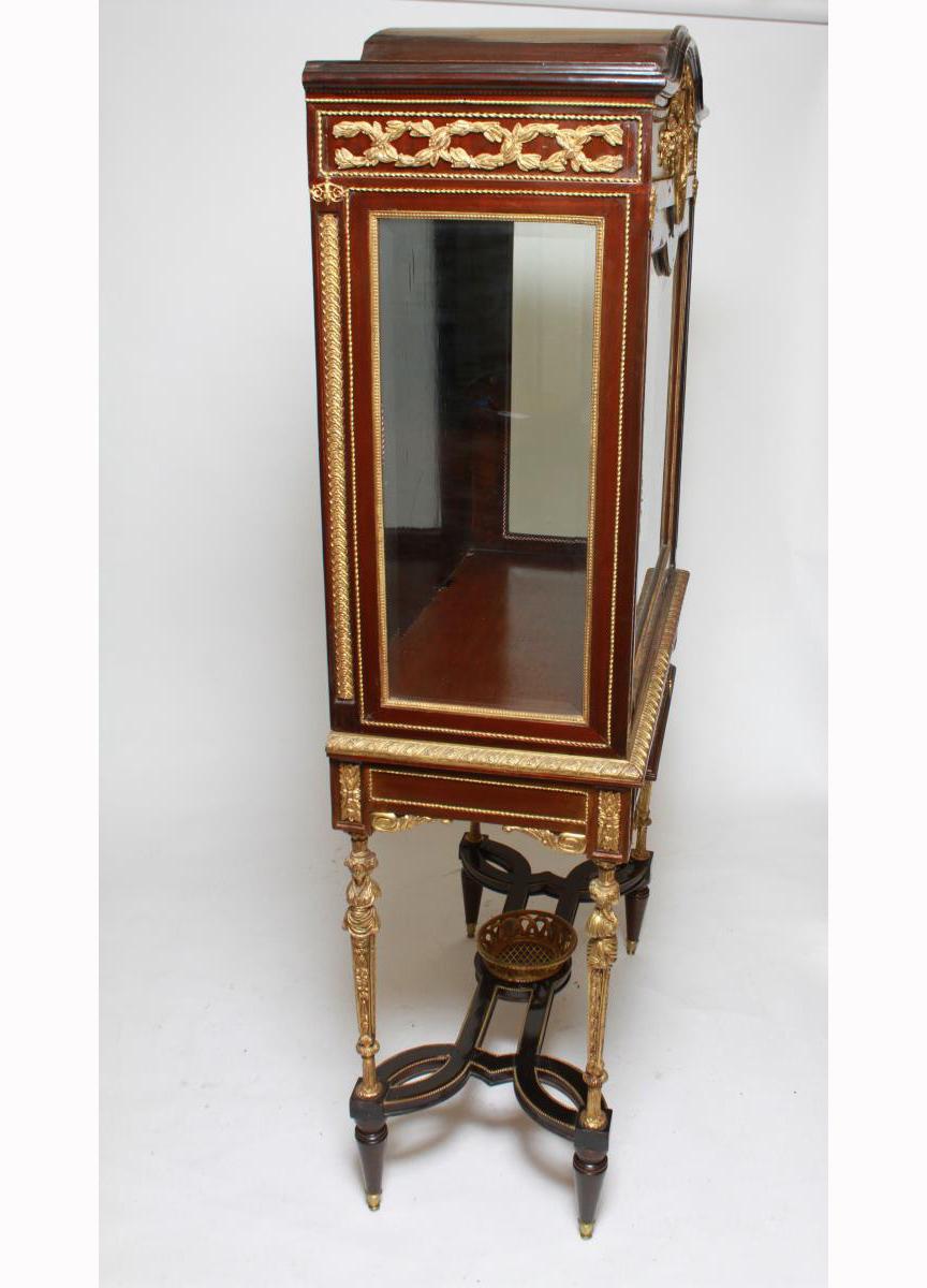 Louis XVI style gilt and ormolu mounted vitrine with panes of beveled glass and a polished ebonized frame with ornate metalwork floral swags, cherubs, mermen, foliage, rope twists and flowers, with a large cartouche enclosing birds. The case is