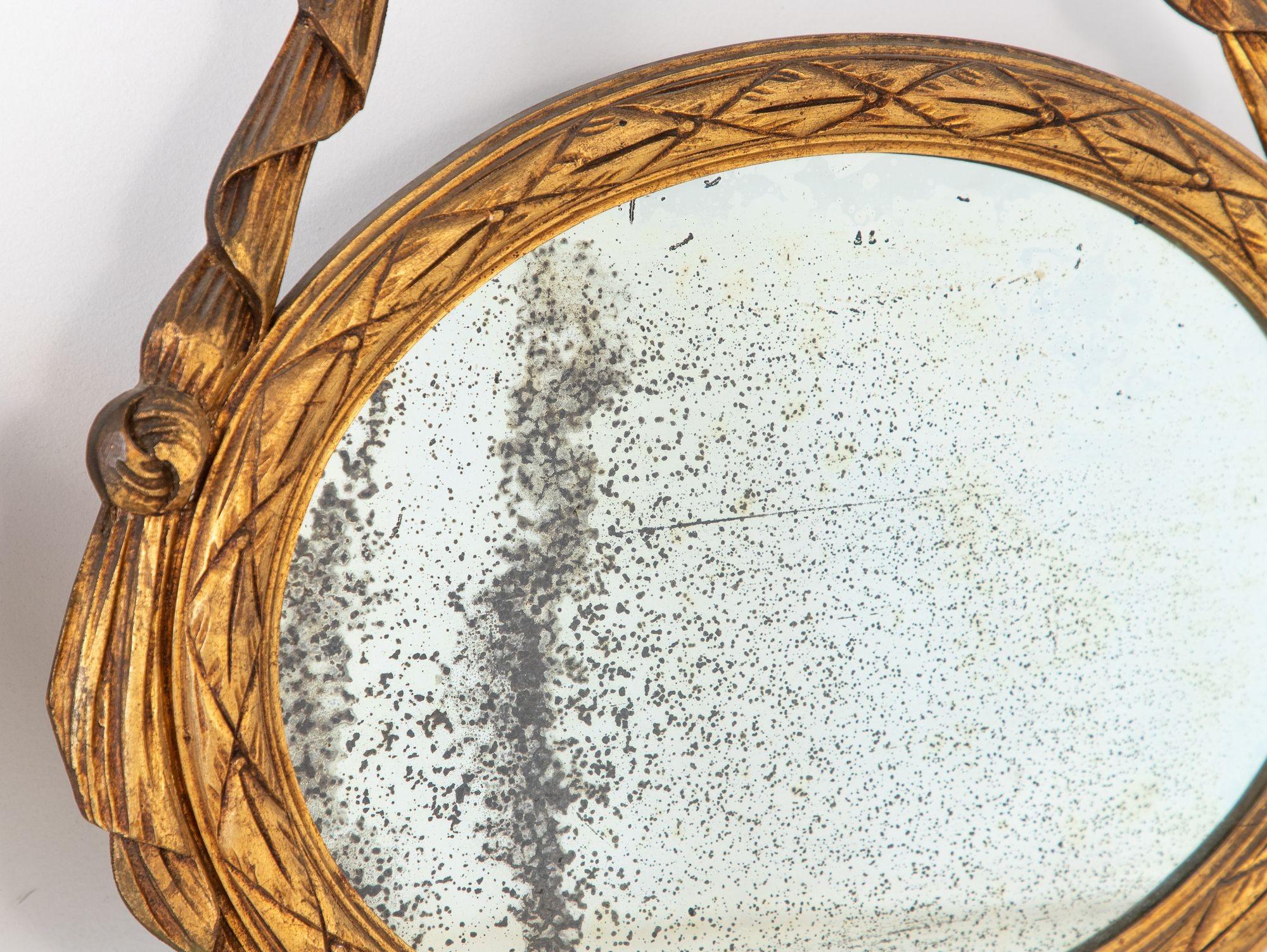 This elegant pair of Louis XVI-style oval mirrors features intricately carved wood frames with floral garlands and classical ribbon crests, all finished in a soft, time-worn gilt. The symmetrical design and refined detailing evoke the grace and