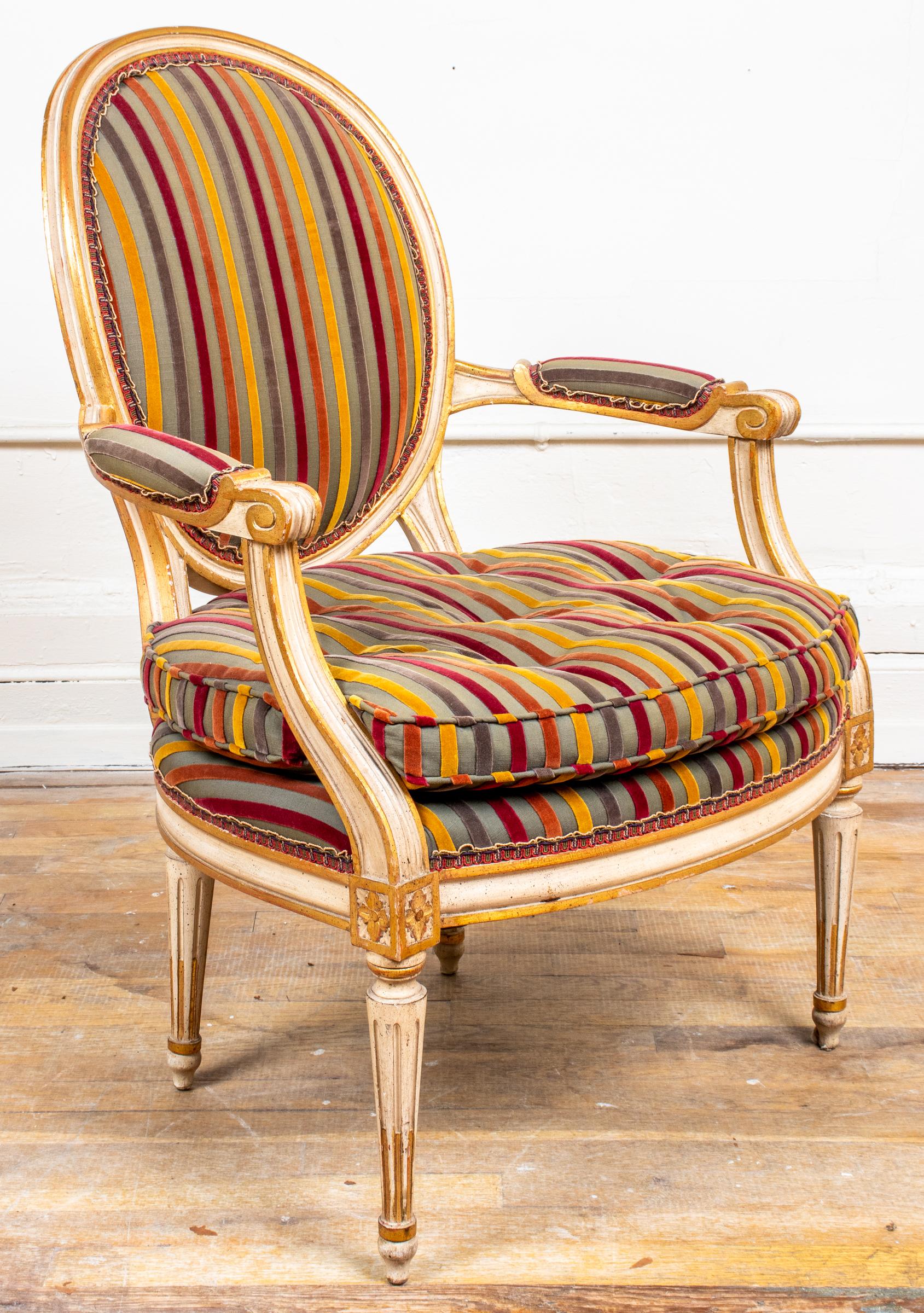 Pair of Louis XVI style fauteuils, the frames parcel gilt and cream painted around the oval backs and cushioned seats with striped velvet upholstery, the scrolling arms with rosette block details above fluted tapered legs. Measures: 35” H x 25” W x