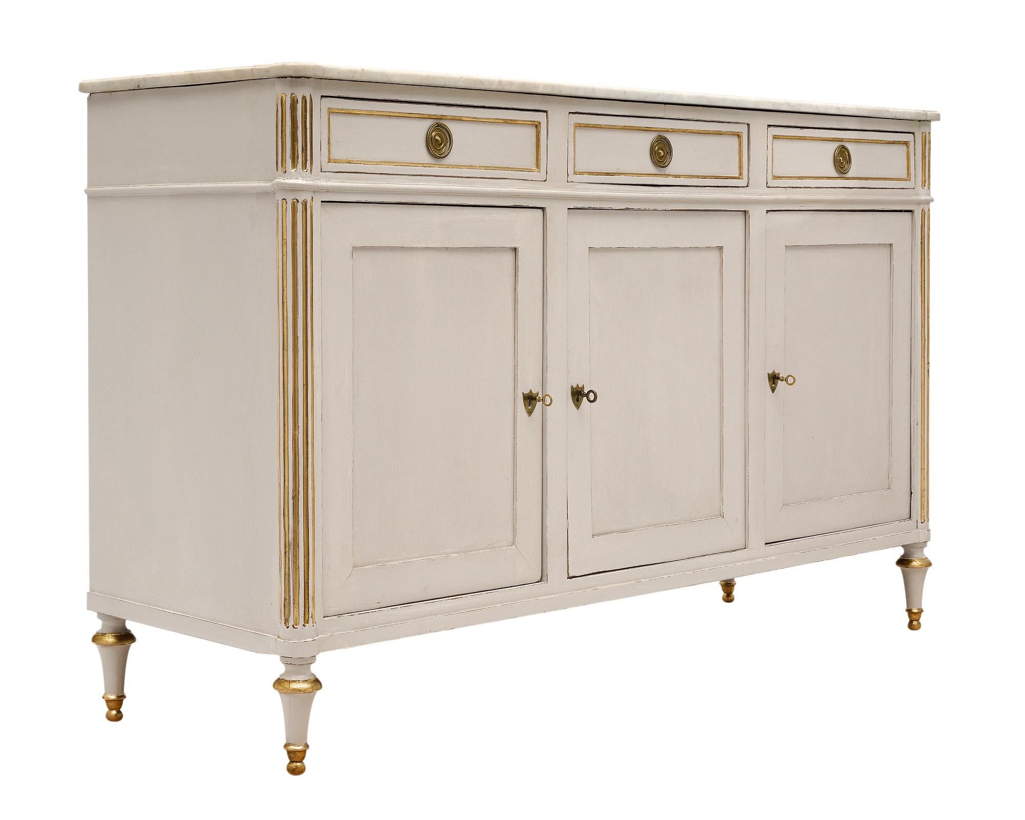 Buffet from France in the Louis XVI style. This case piece has been painted in a cream colored milk paint with 23 carat gold leaf in the fluted detailing and feet. There are three dovetailed drawers and three doors all with original, finely cast