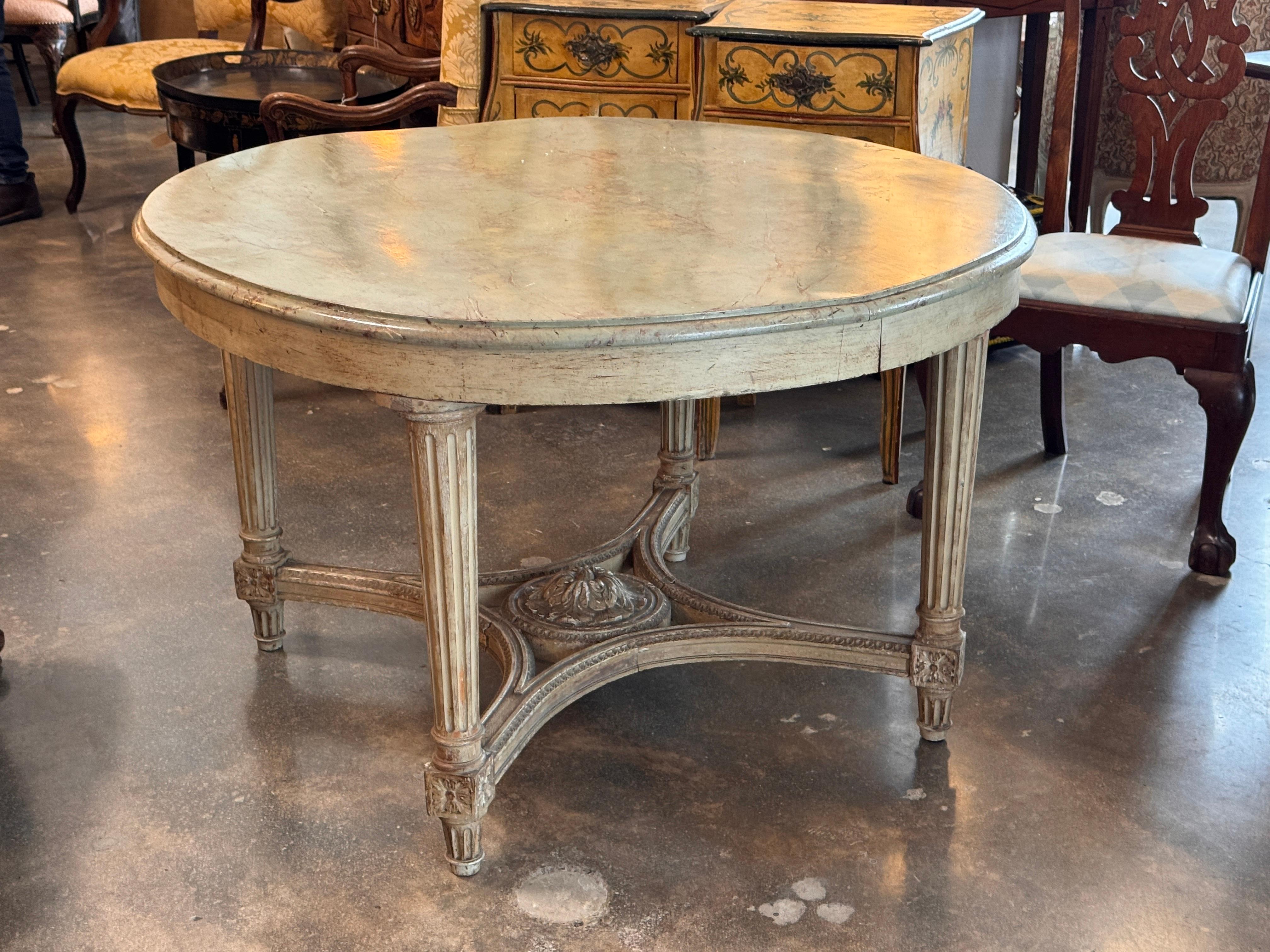Center table in the Louis XVI style with a painted and faux-marble top over fluted legs joined by an arched X-stretcher. Carved rosettes and a central carved medallion highlight the base, and the painted surface shows the aged texture visible in the
