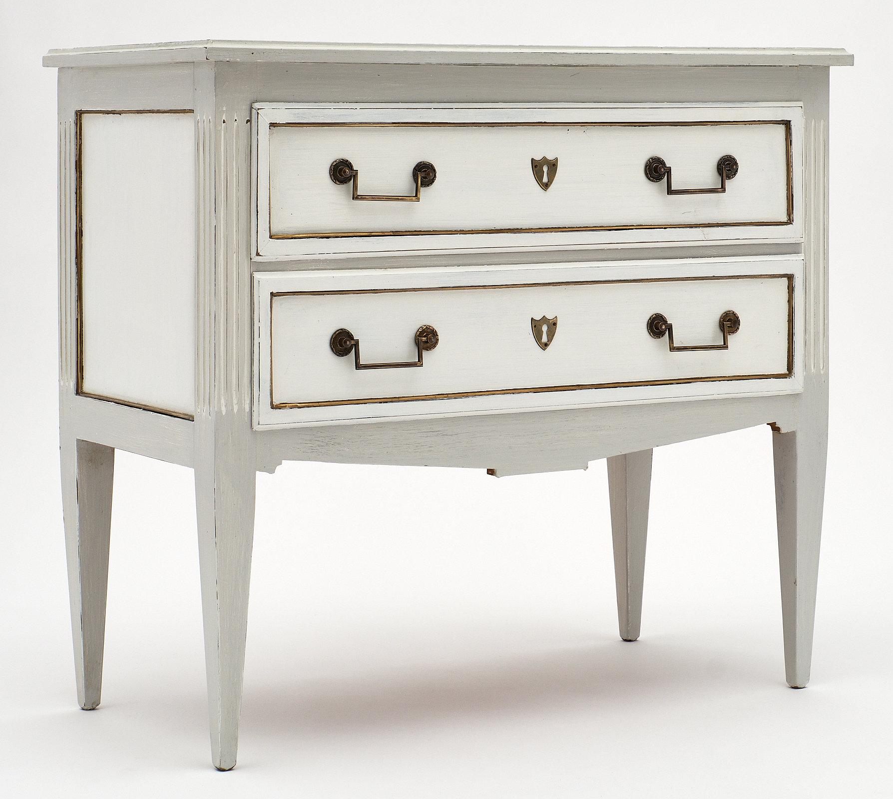 Painted Louis XVI style chest with brass trim. This elegant, petite chest of drawers “commode sauteuse” is made of cherry wood and features a “Trianon” gray and white patina, brass hardware and trims. Two large dovetailed drawers offer ample