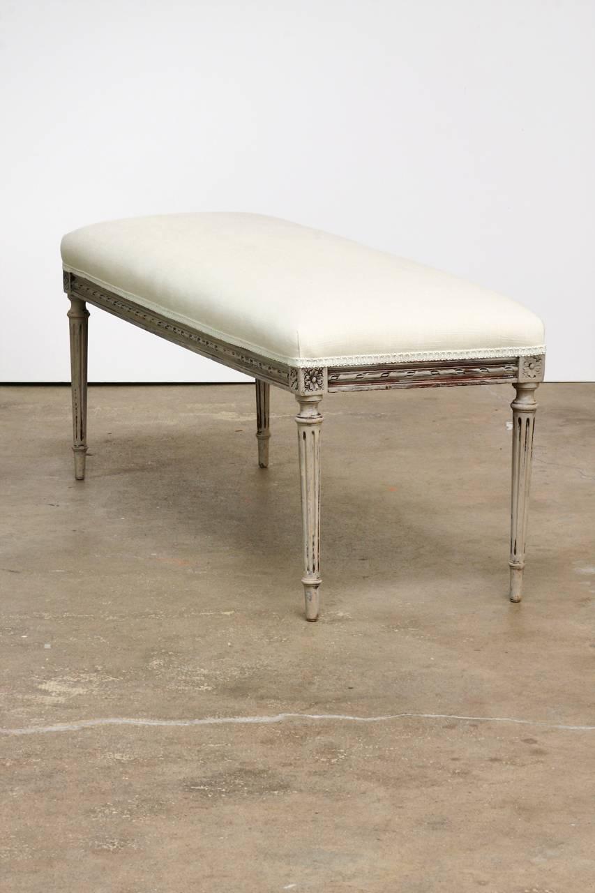 Charming painted bench made in the Louis XVI taste. Newly upholstered in French linen with a Gustavian style painted finish of grey. The carved frame features rosettes on the corners and elegant tapered, fluted legs. The bench has been