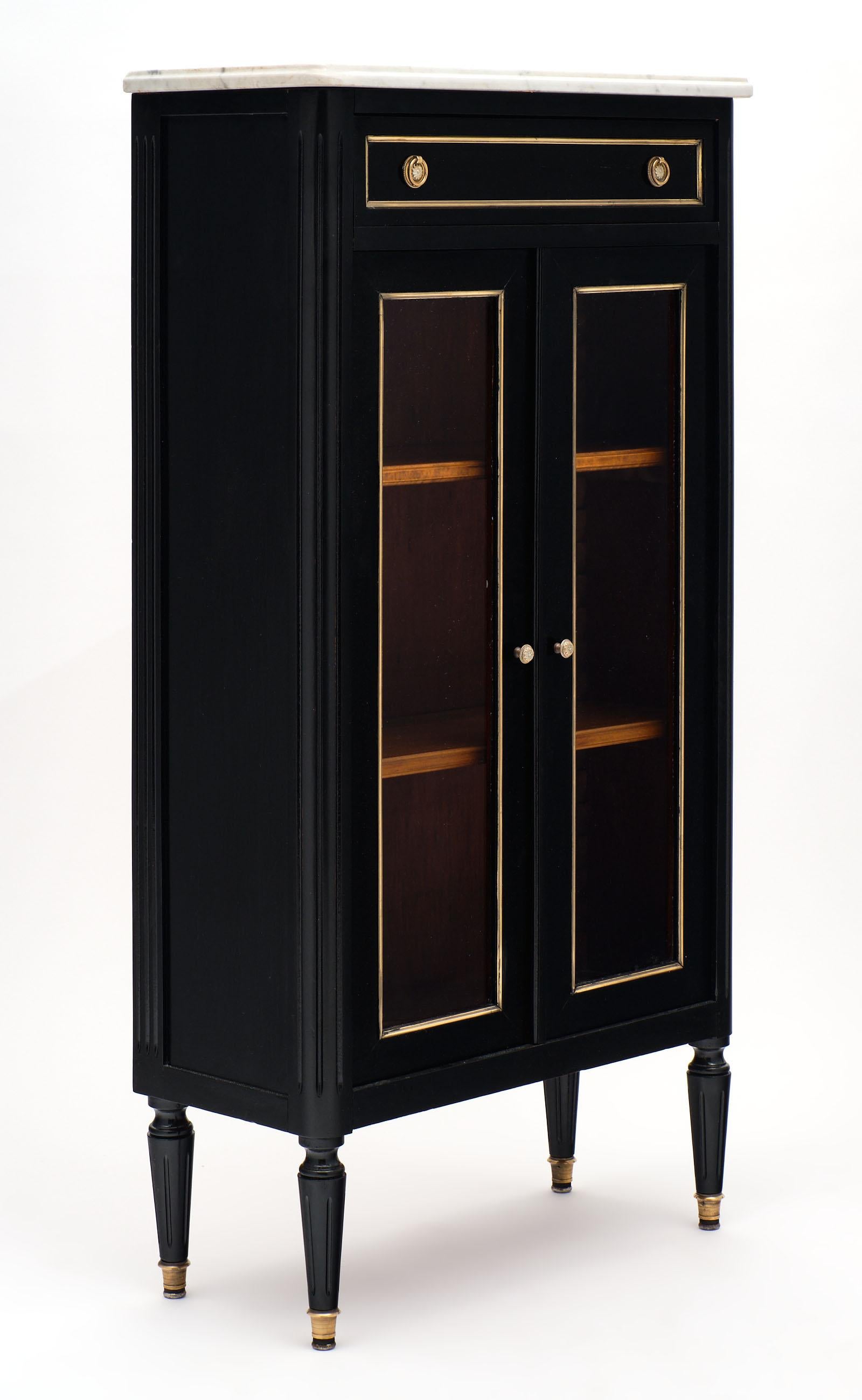 Petite ebonized Louis XVI style bookcase with Classic French elegance and two glass doors trimmed in brass. There is also a dovetailed top drawer and a beautiful Carrara marble top. This piece is made of mahogany that has been finished with a French