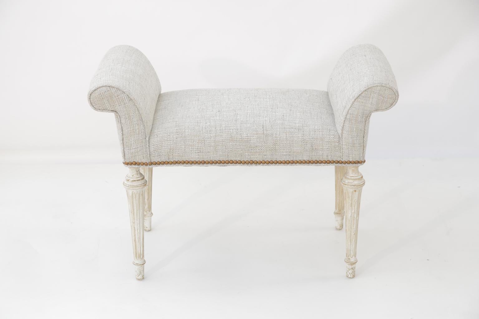 Bench, having generous rolled arms flanking its crown seat, its ivory painted base showing natural wear; raised on four, round, fluted, tapering legs, ending in touipe feet. Upholstered in a light blue weave with nailheads.

Stock ID: D3489