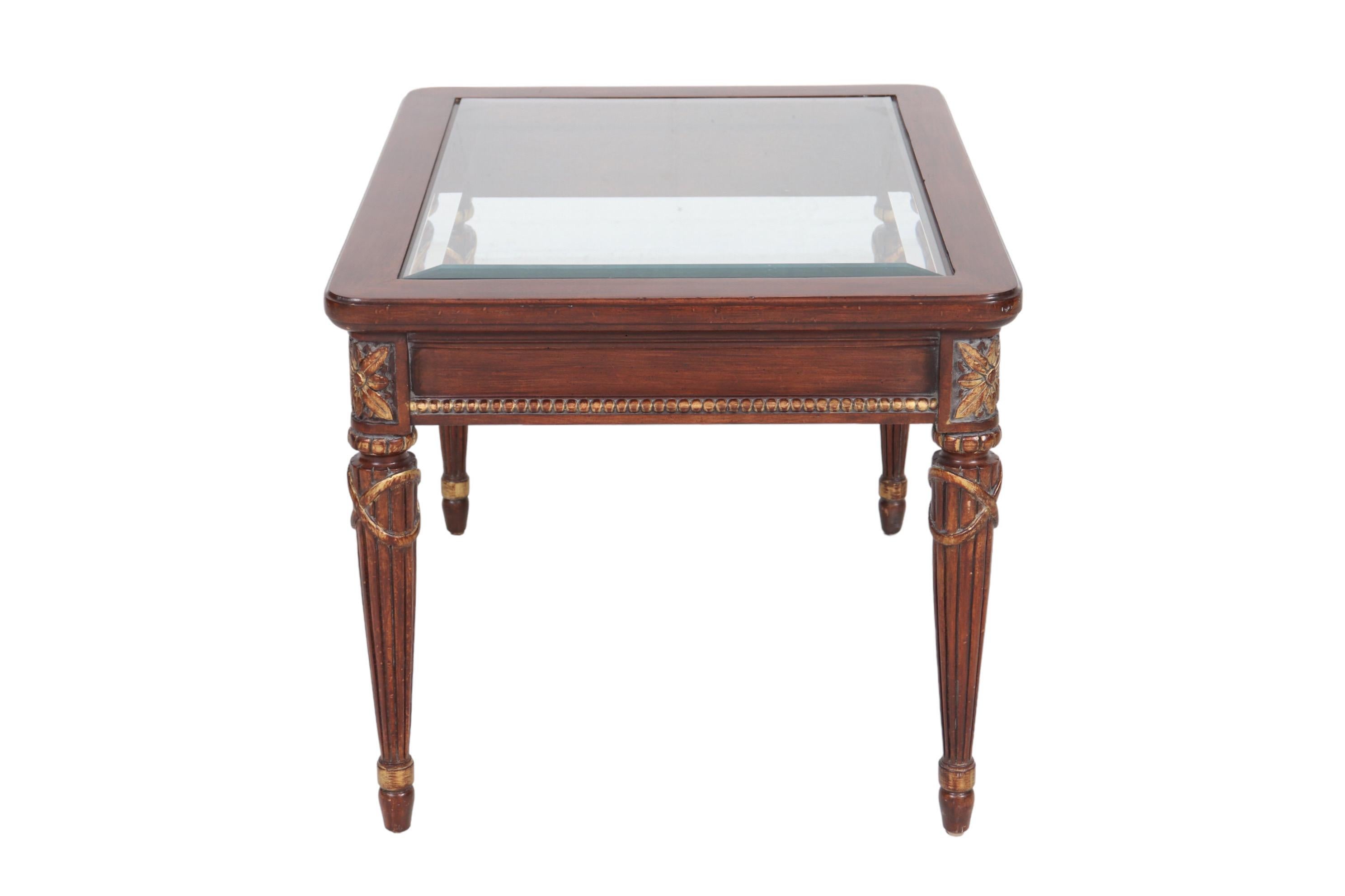 Table d'appoint de style Louis XVI avec un plateau en verre biseauté. La jupe de la table est ornée d'une bordure de perles sculptées. Des rosettes sculptées surmontent des pieds ronds et cannelés qui s'affinent et se terminent par des pieds en