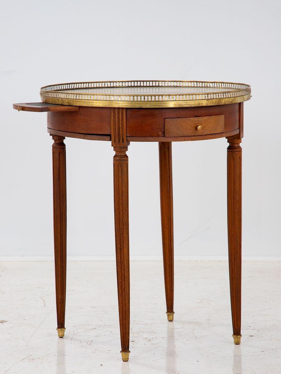 A round Late 19th century Louis XVI style bouillotte table with a marble top, brass gallery, and brass capped feet. Mahogany veneer with tapered legs. This side table has a single drawer. Minor loss to veneer.