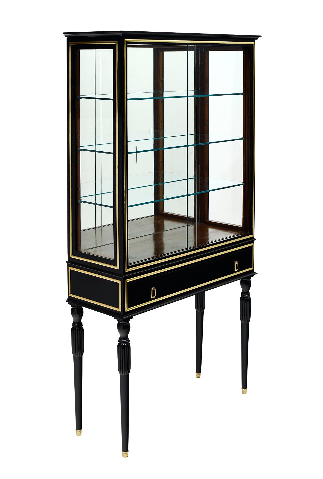 This French Louis XVI Vitrine is made of Mahogany ebonized and finished in a high luster Museum quality French polish. The cabinet is trimmed in gilt brass and features a dovetailed drawer; a back mirror; three glass shelves and clear glass sides.