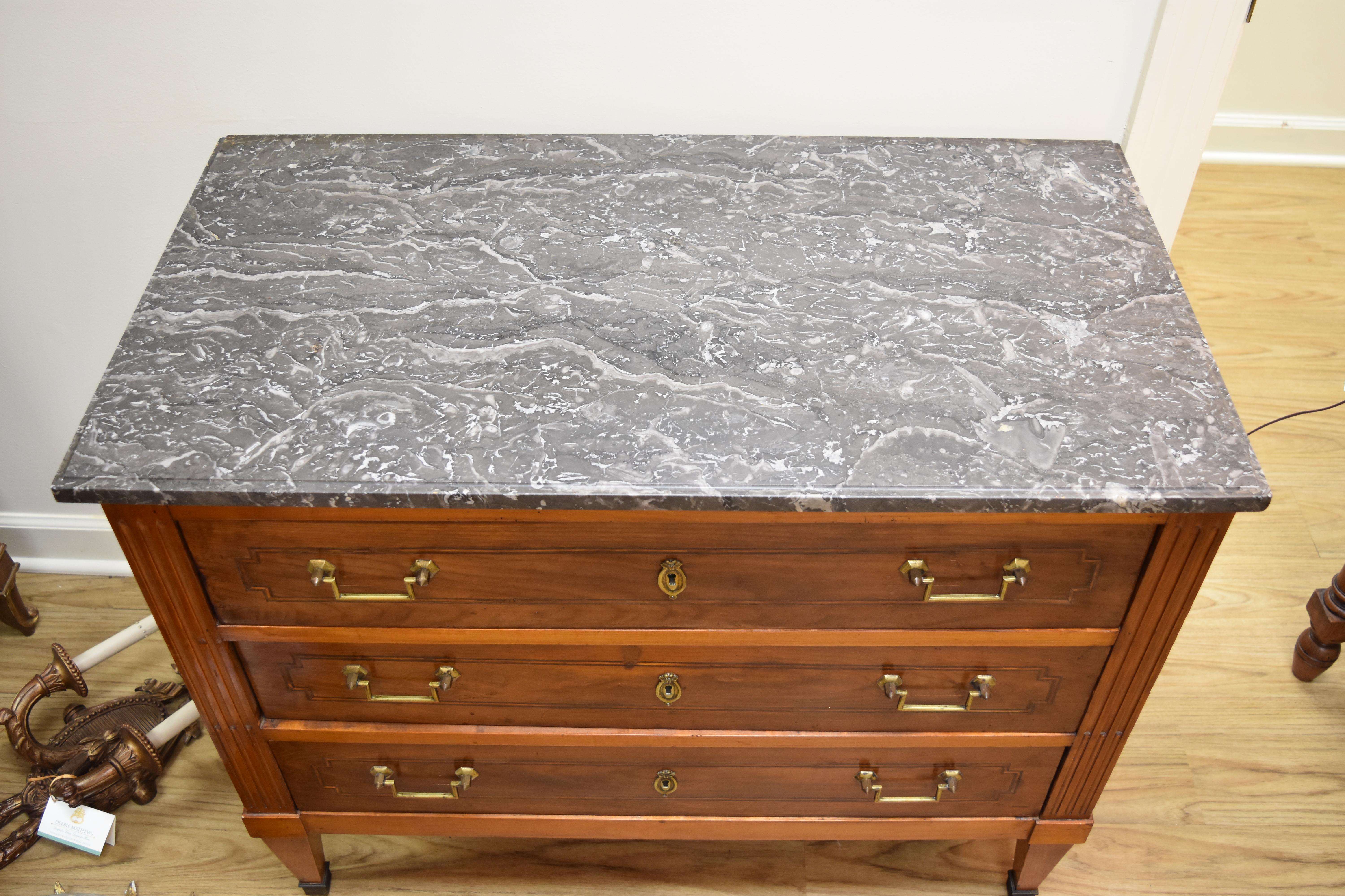 This Louis XVI style walnut chest features three working drawers with a geometric patterned inlay design. The drawers have their original bronze drop pulls and key escutcheons. The gray marble top is beveled around the edges and rests on fluted side