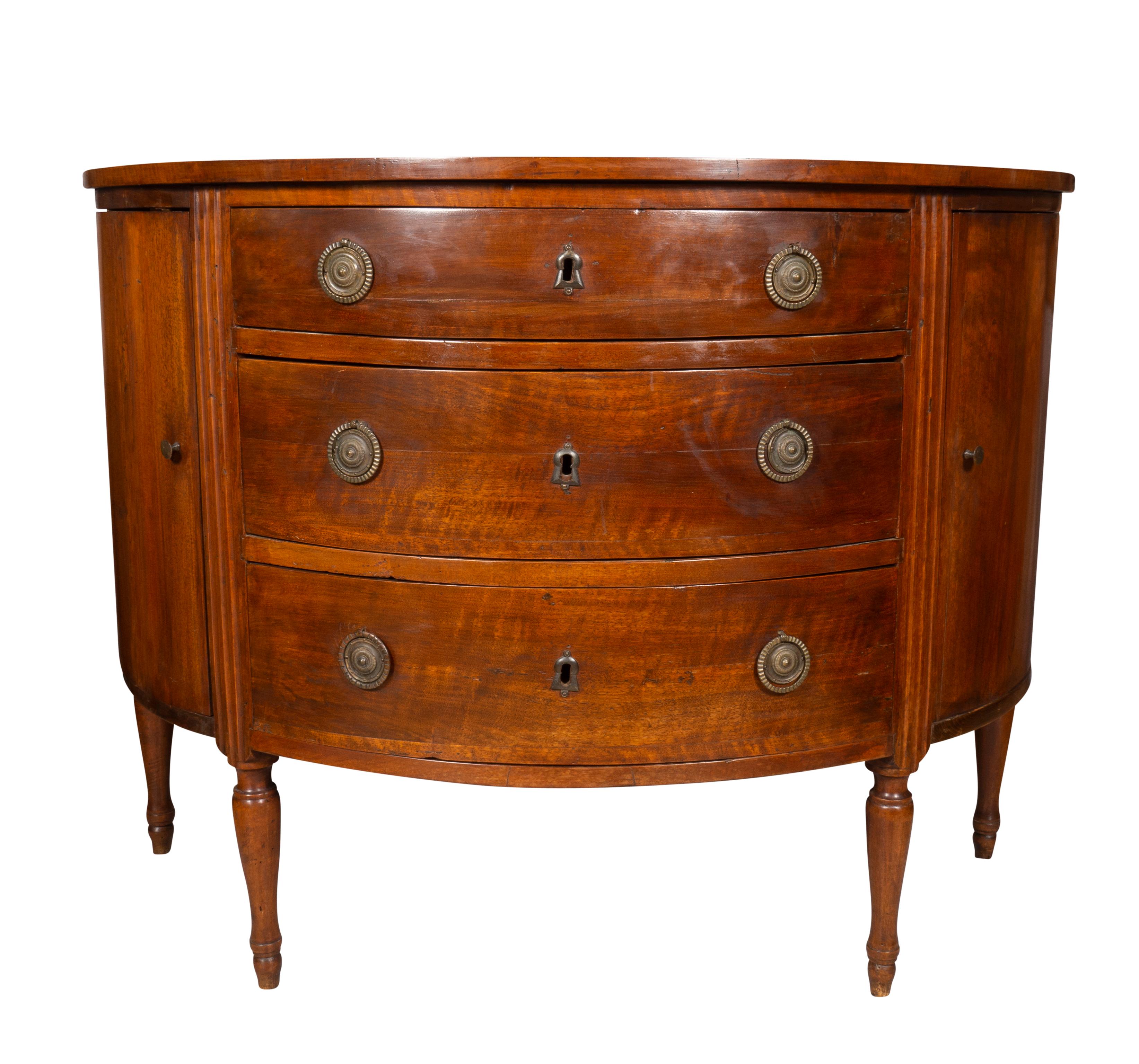 A hard to find form. With a demilune top over a conforming case with three drawers with brass ring handles and round backplates flanked by fluted columns and a pair of cabinet doors enclosing shelves, raised on circular tapered legs.