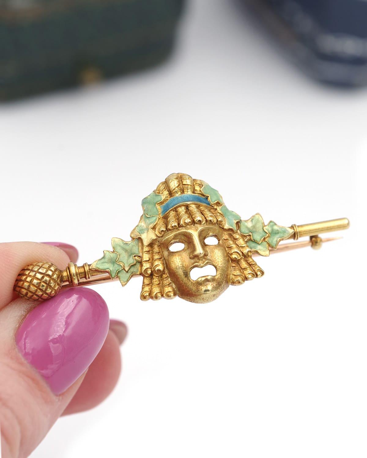 Broche Art Nouveau de máscara teatral Louis Zorra en oro de 18 quilates, con una antigua máscara teatral griega en oro mate, rematada con una cinta esmaltada en azul y hojas de hiedra esmaltadas en verde. El broche barette termina en un motivo de