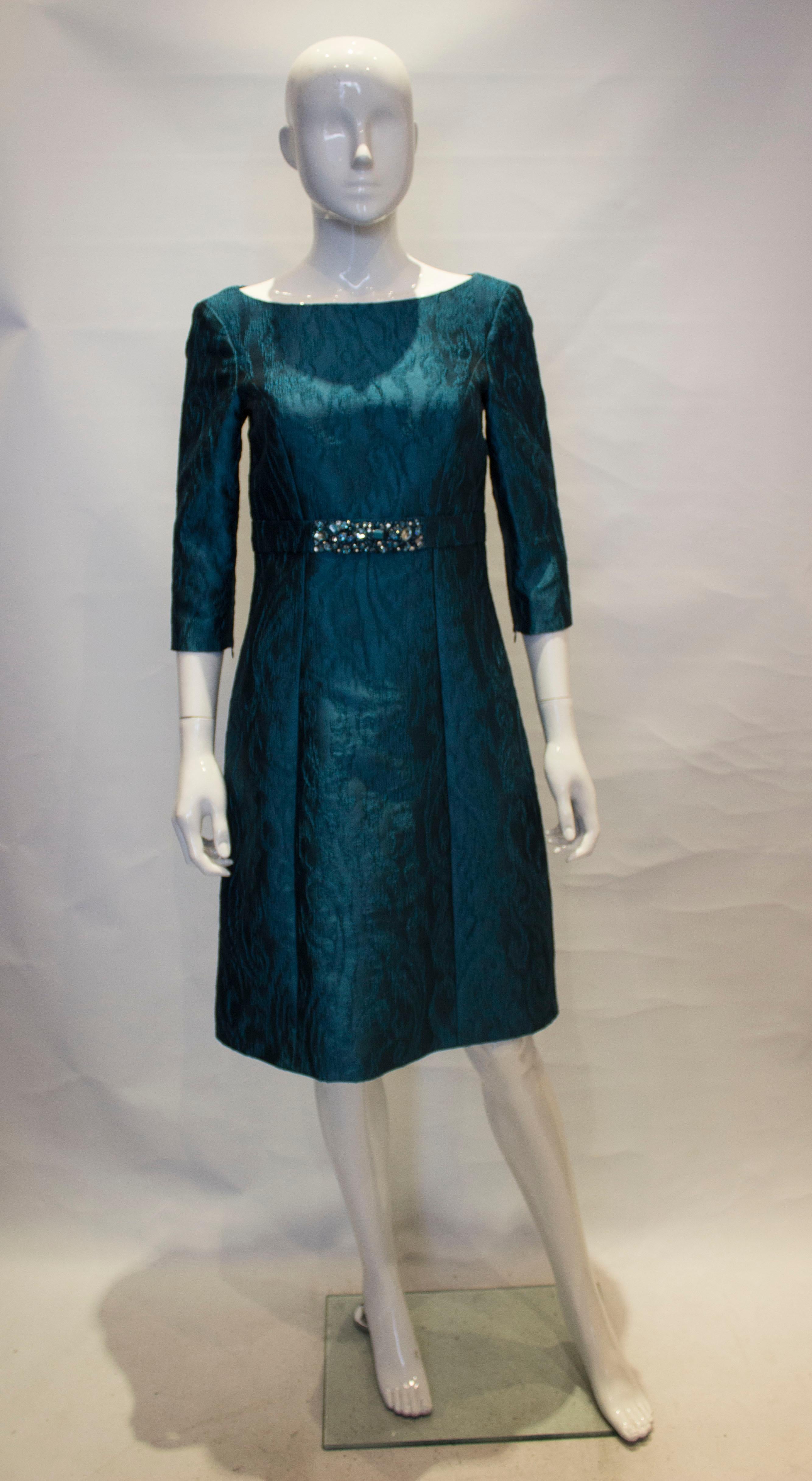 A chic cocktail dress by Louisa Spagnoli. The dress is in a vibrant blue in a textured fabric, and is fully lined. It has a central back zip, with zip on the elbow length sleaves, and a decorated belt at waist leval.
